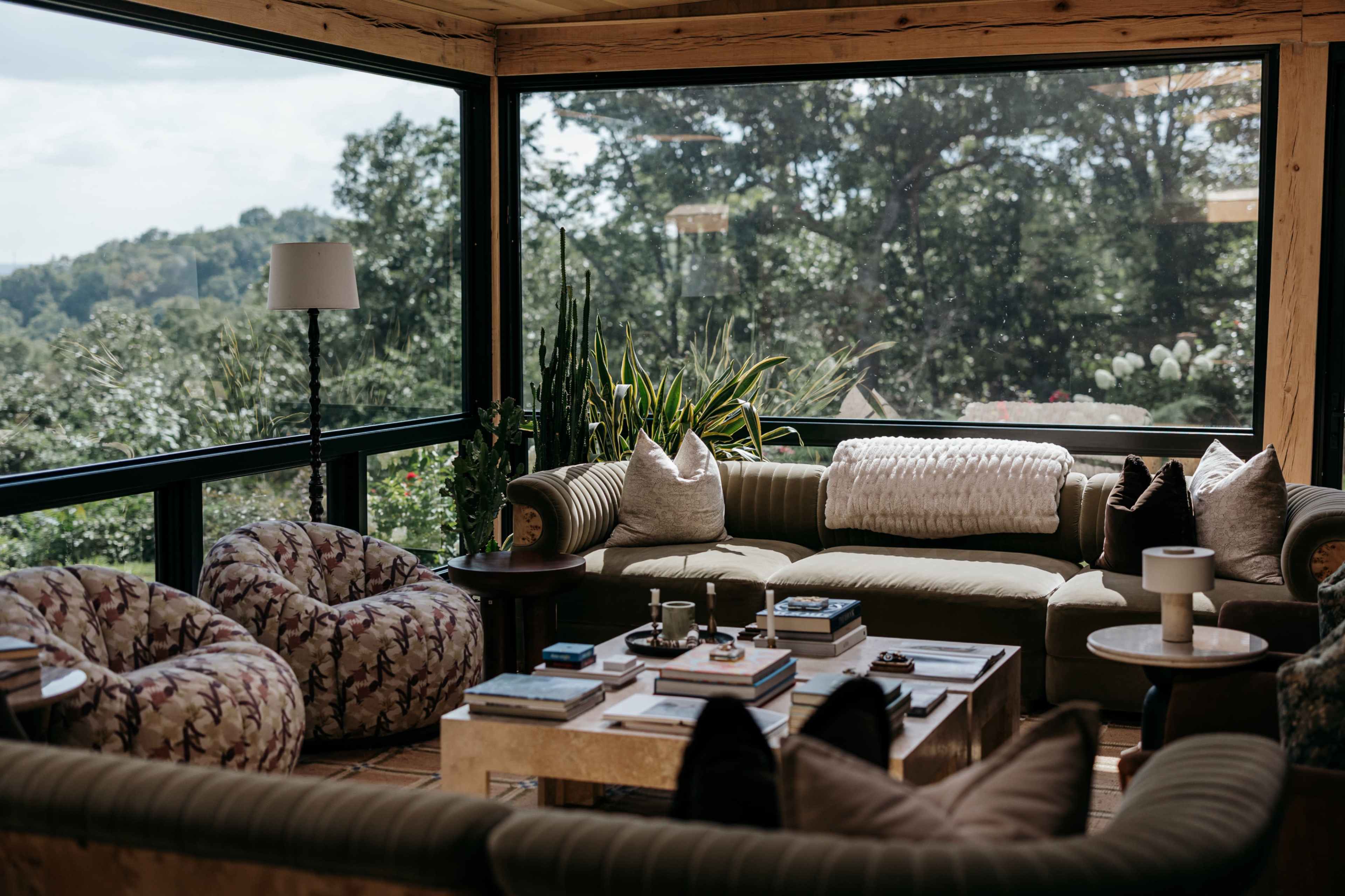 The image shows a modern living room furnished with a large green sectional sofa, two patterned bean bag chairs, a coffee table stacked with books, and large windows offering a view of greenery outside.