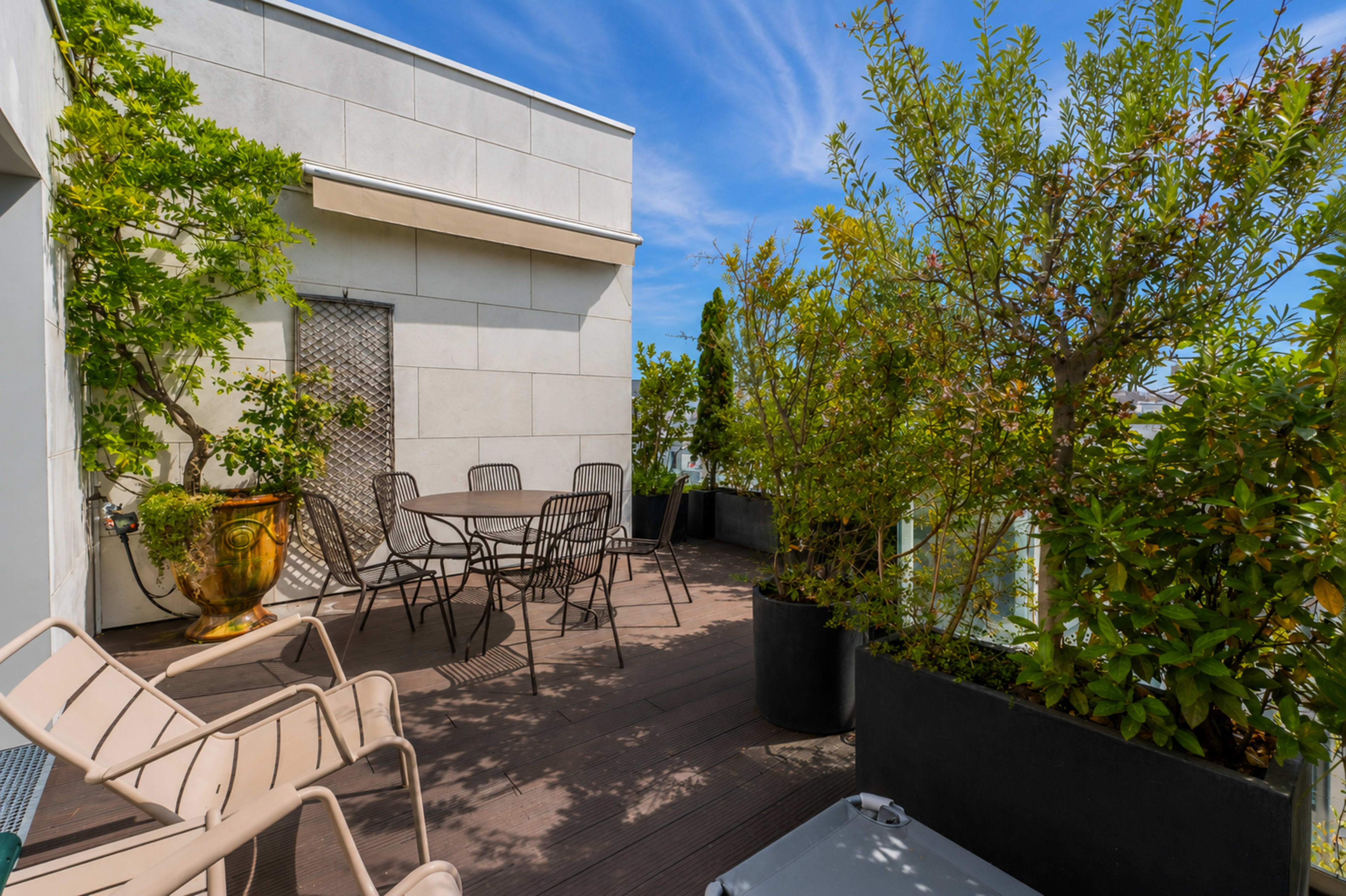 The image shows a rooftop terrace decorated with potted plants, a small table and chairs, and a blue sky overhead.