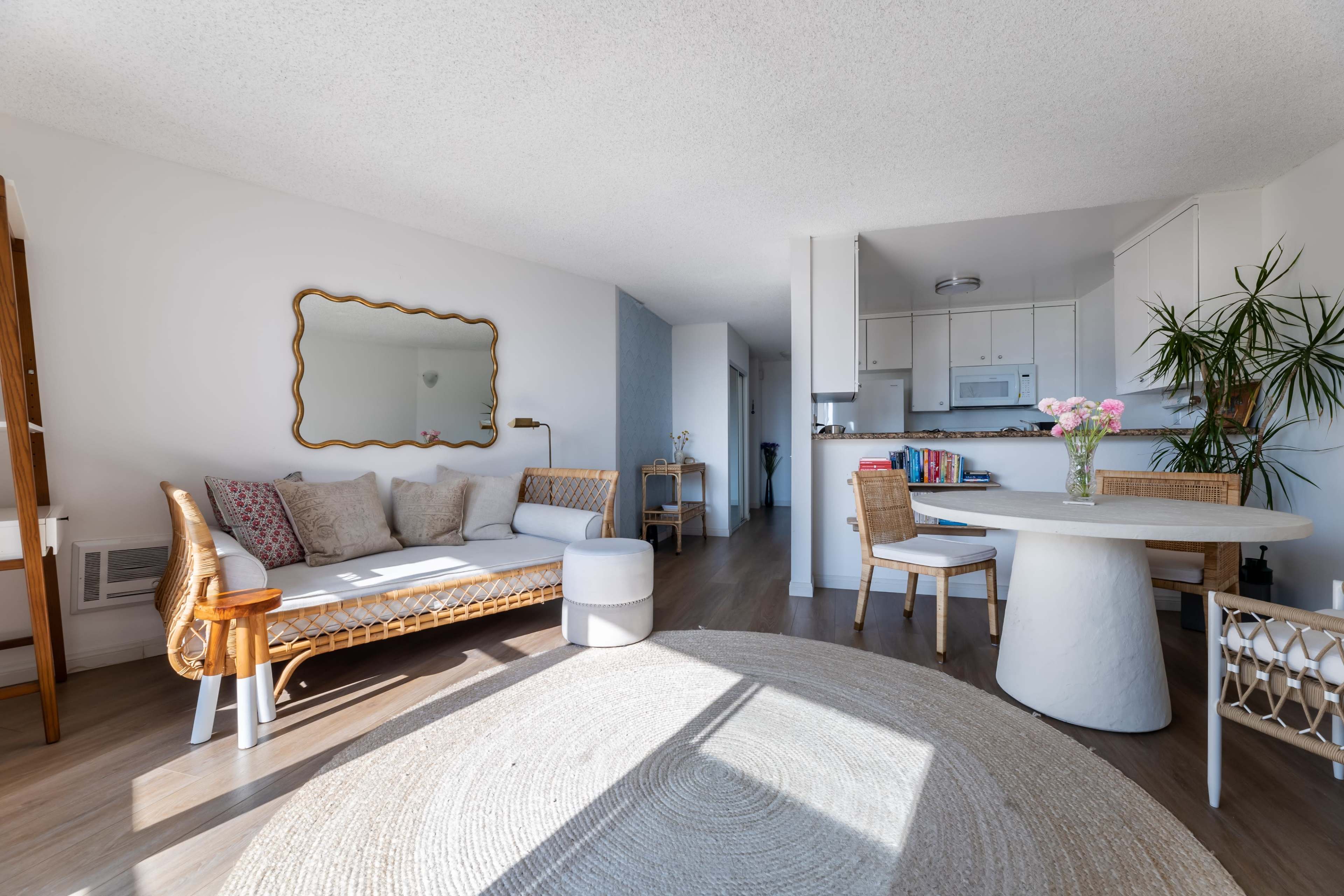 The image depicts a bright and modern living space featuring a rattan sofa, a round dining table with chairs, and a large area rug.