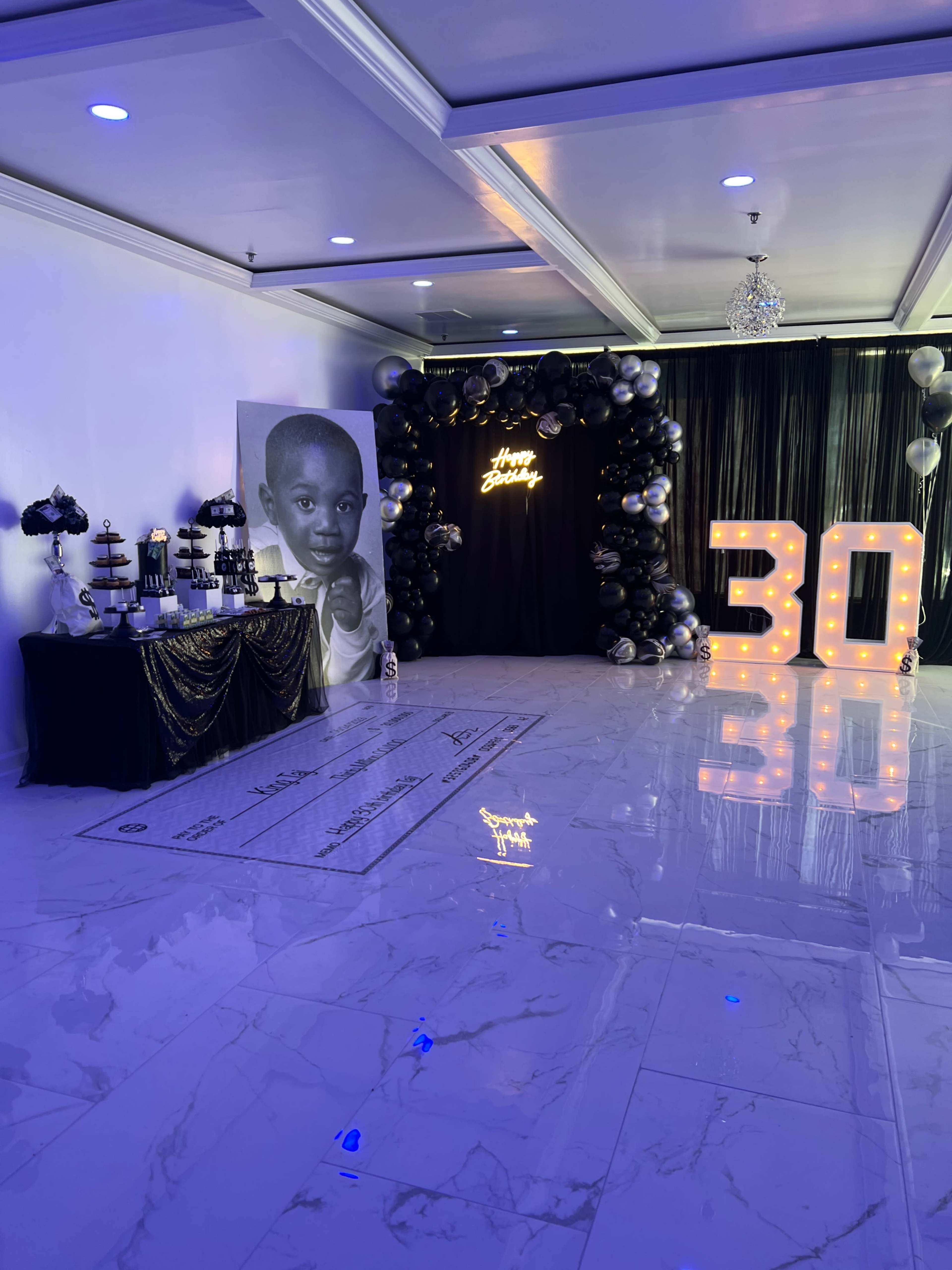 A decorated indoor venue for a 30th birthday celebration, featuring a dessert table, a large black and white portrait, and illuminated "30" balloons.