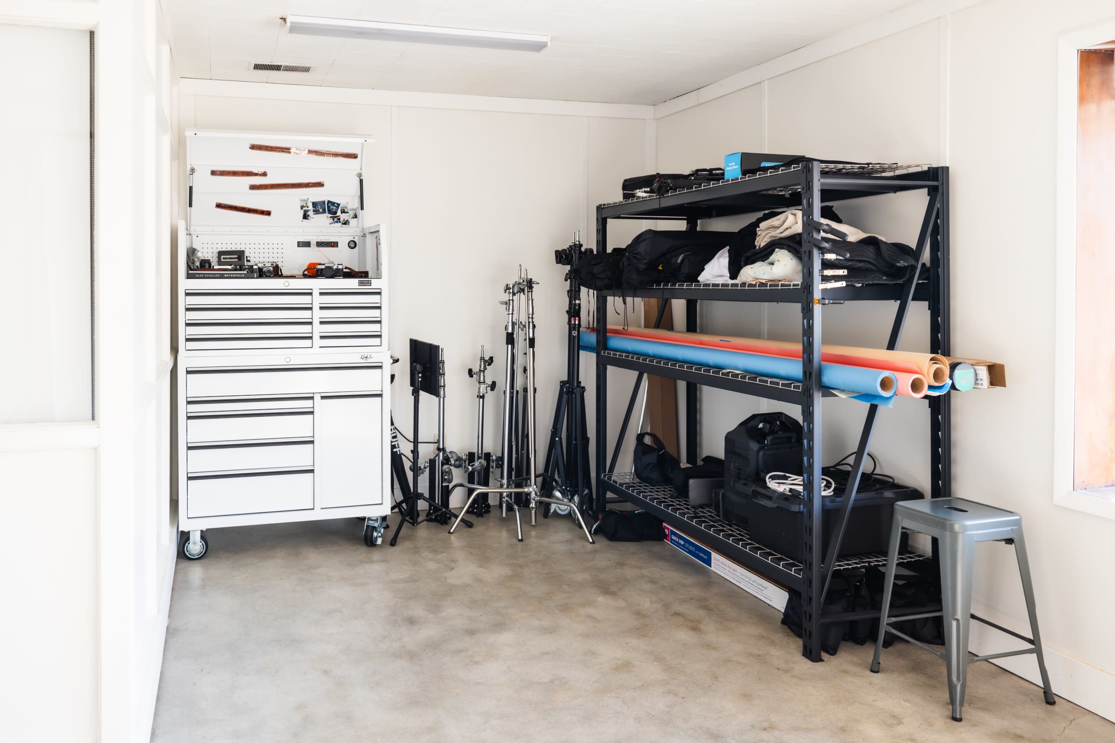 The image shows a storage room with a metal tool chest on wheels, various photography equipment on stands, and shelves holding rolls of paper and additional gear.