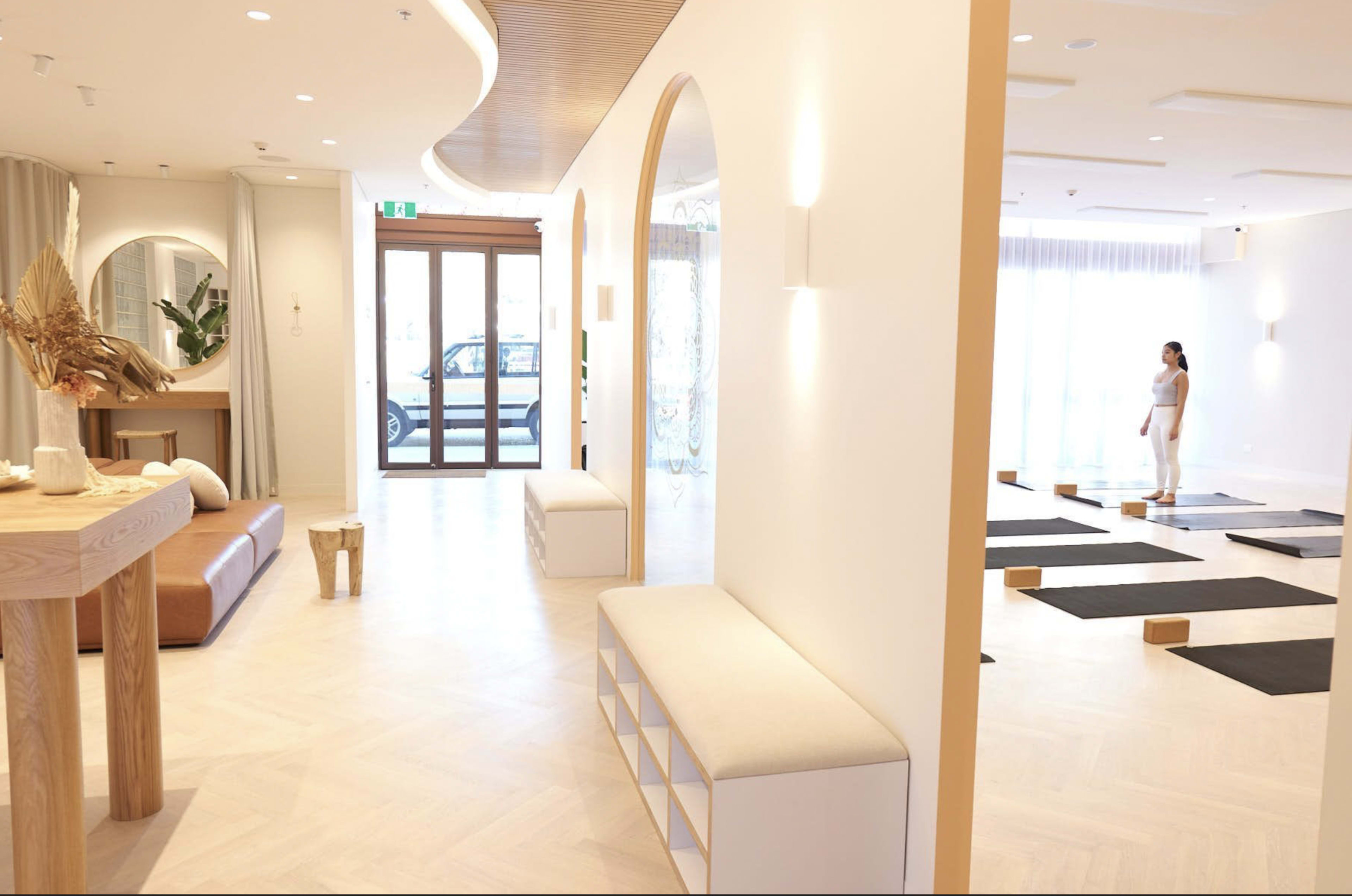 A modern, well-lit yoga studio with a minimalist interior, featuring a seating area, a large mirror, and yoga mats arranged on the floor.