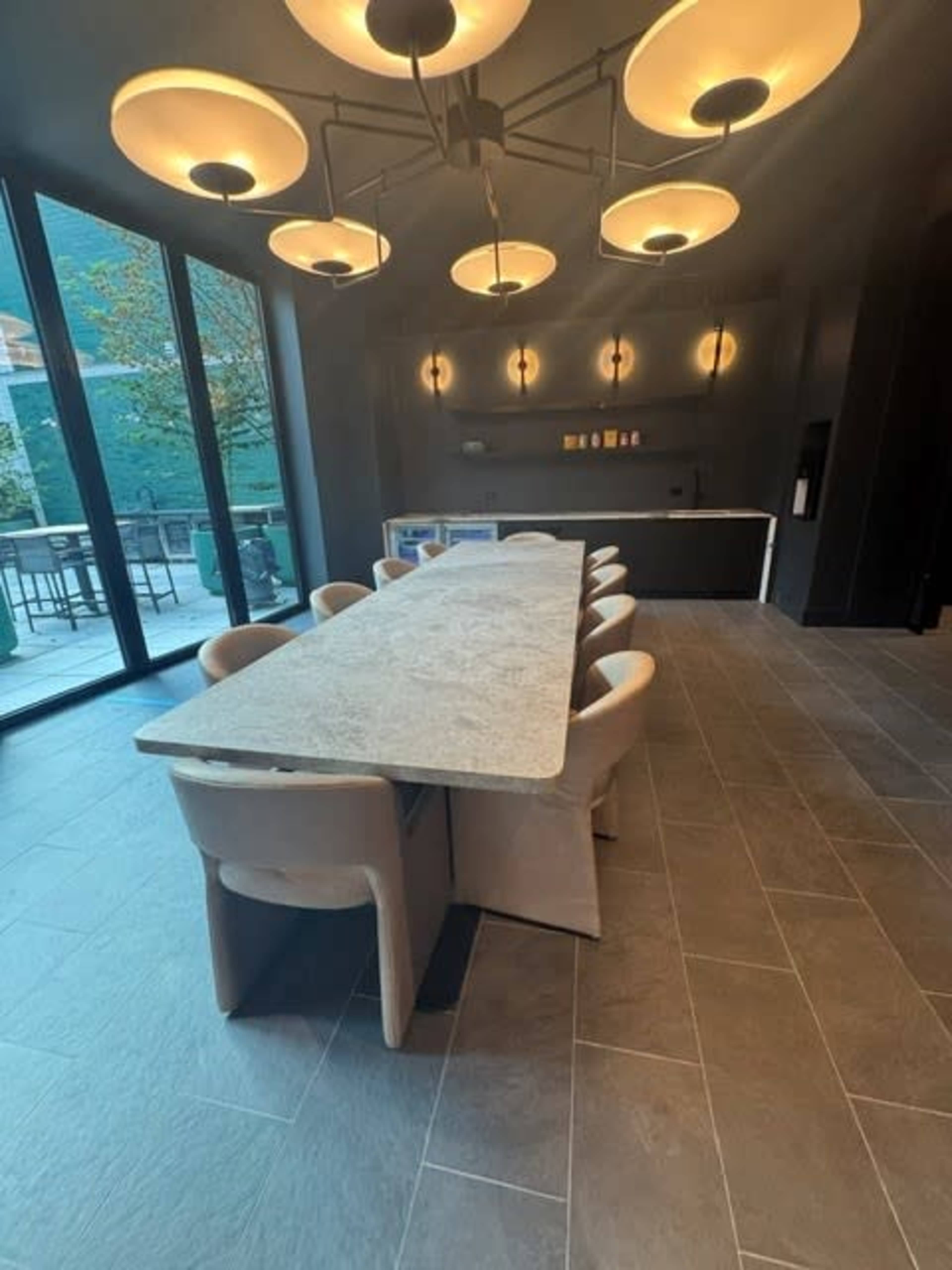 The image shows a spacious dining area featuring a long, rectangular table surrounded by beige chairs, beneath modern pendant lights.