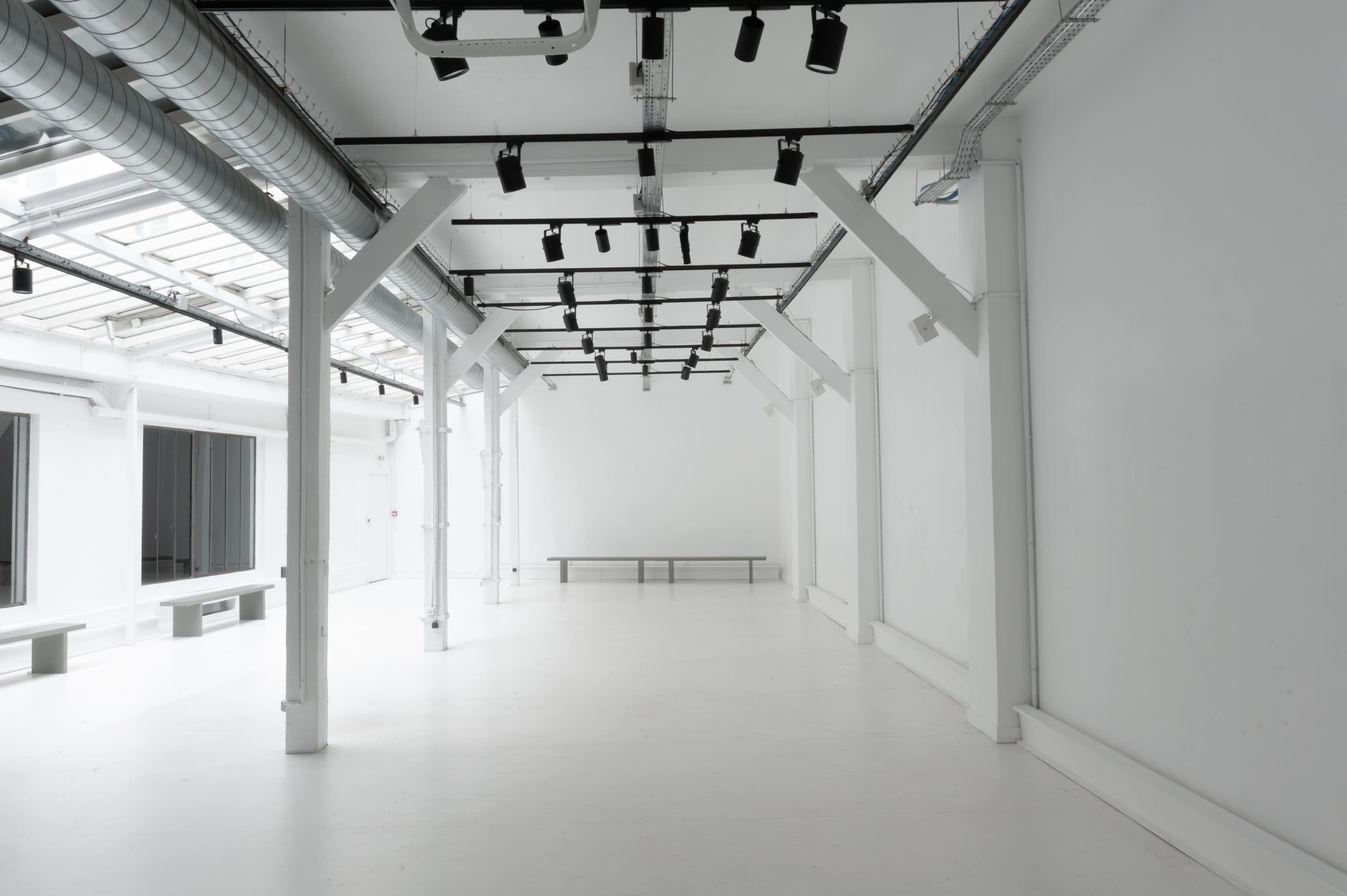 The image shows a large, empty, and minimalist white room with overhead lighting and a simple bench along one wall.