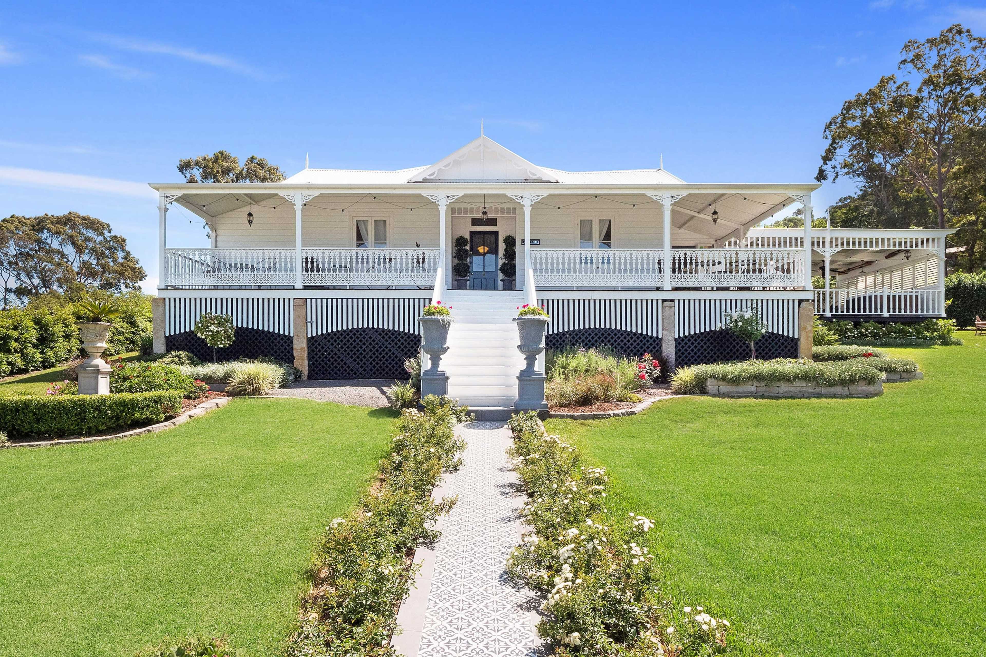 A large, white wooden house with a verandah sits on a manicured lawn, surrounded by landscaped gardens and a patterned path leading to the entrance.