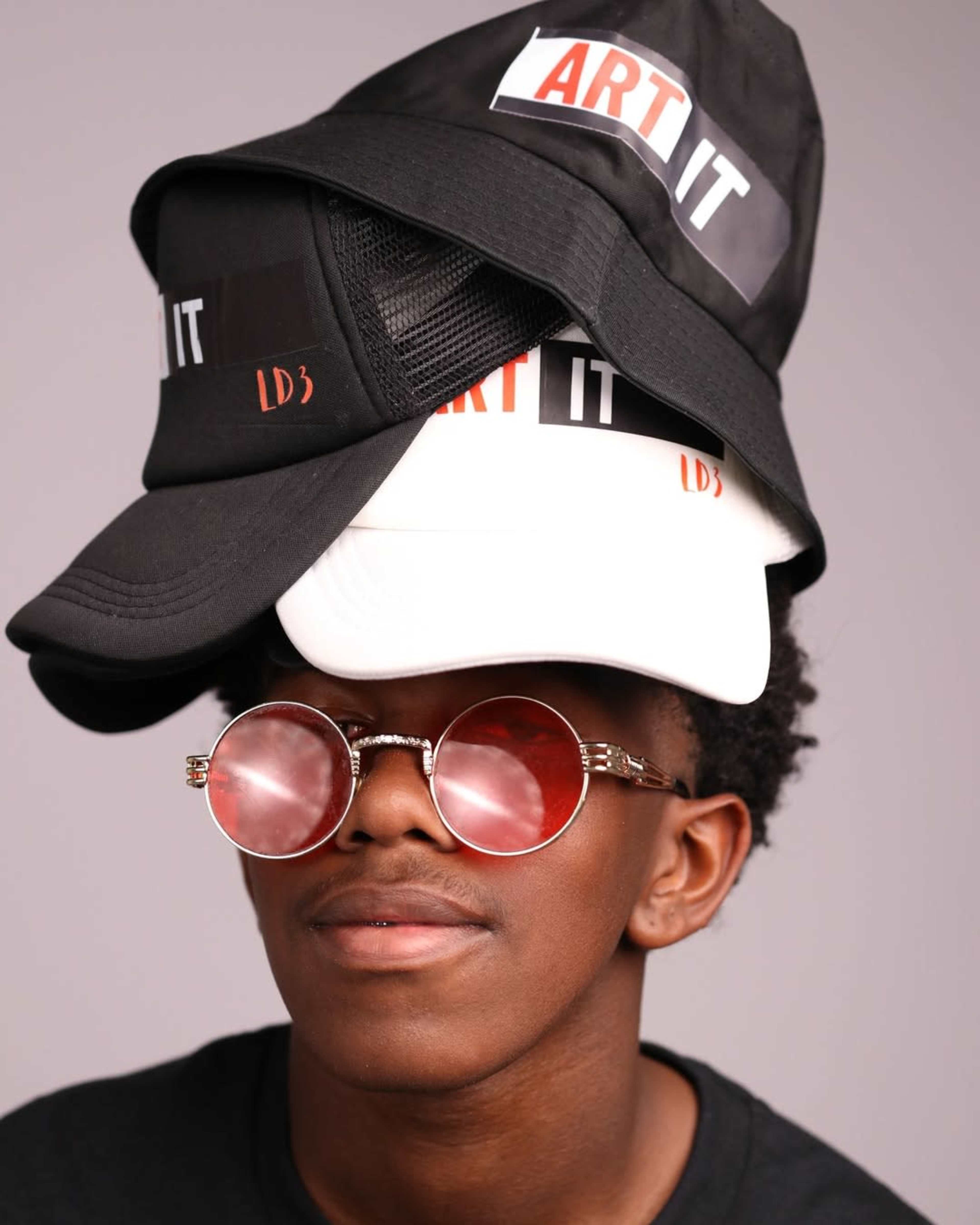 A person is wearing multiple hats stacked on their head and round sunglasses, posing against a neutral background.
