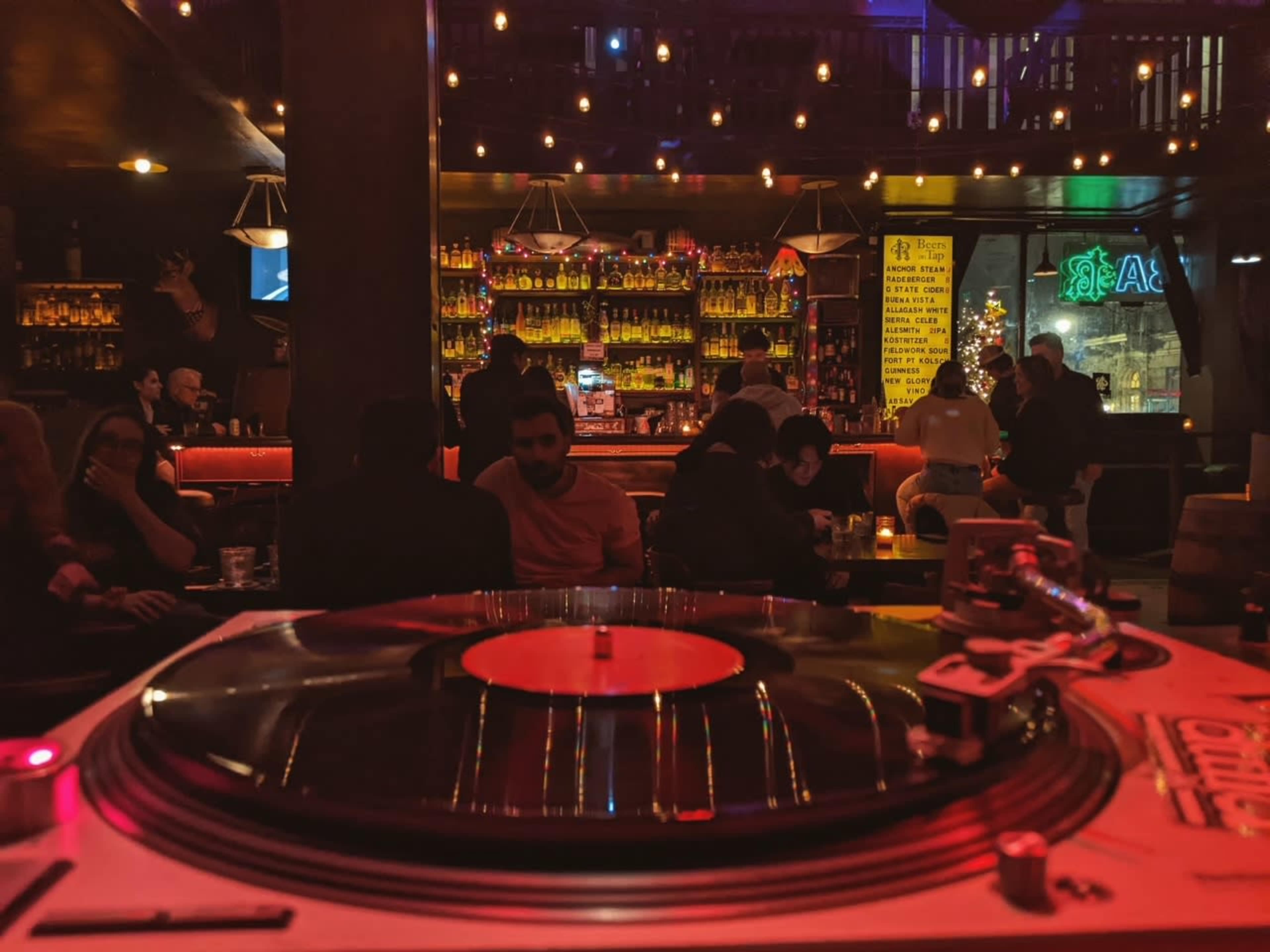 A vinyl record player is in the foreground, while a bar filled with patrons and illuminated bottles is visible in the background.