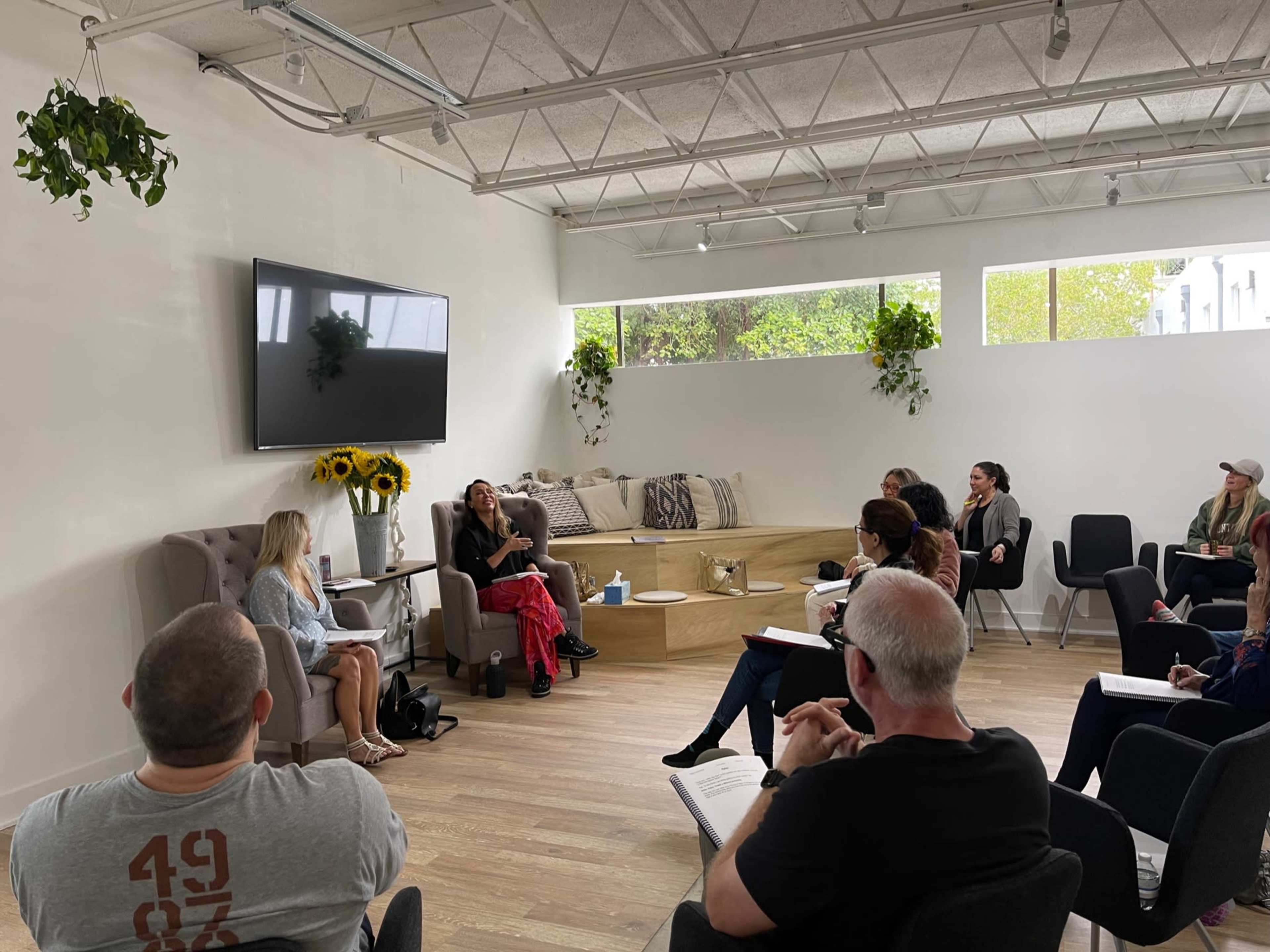 A group of people sits in a modern, well-lit room, engaged in a discussion with two individuals on a stage.