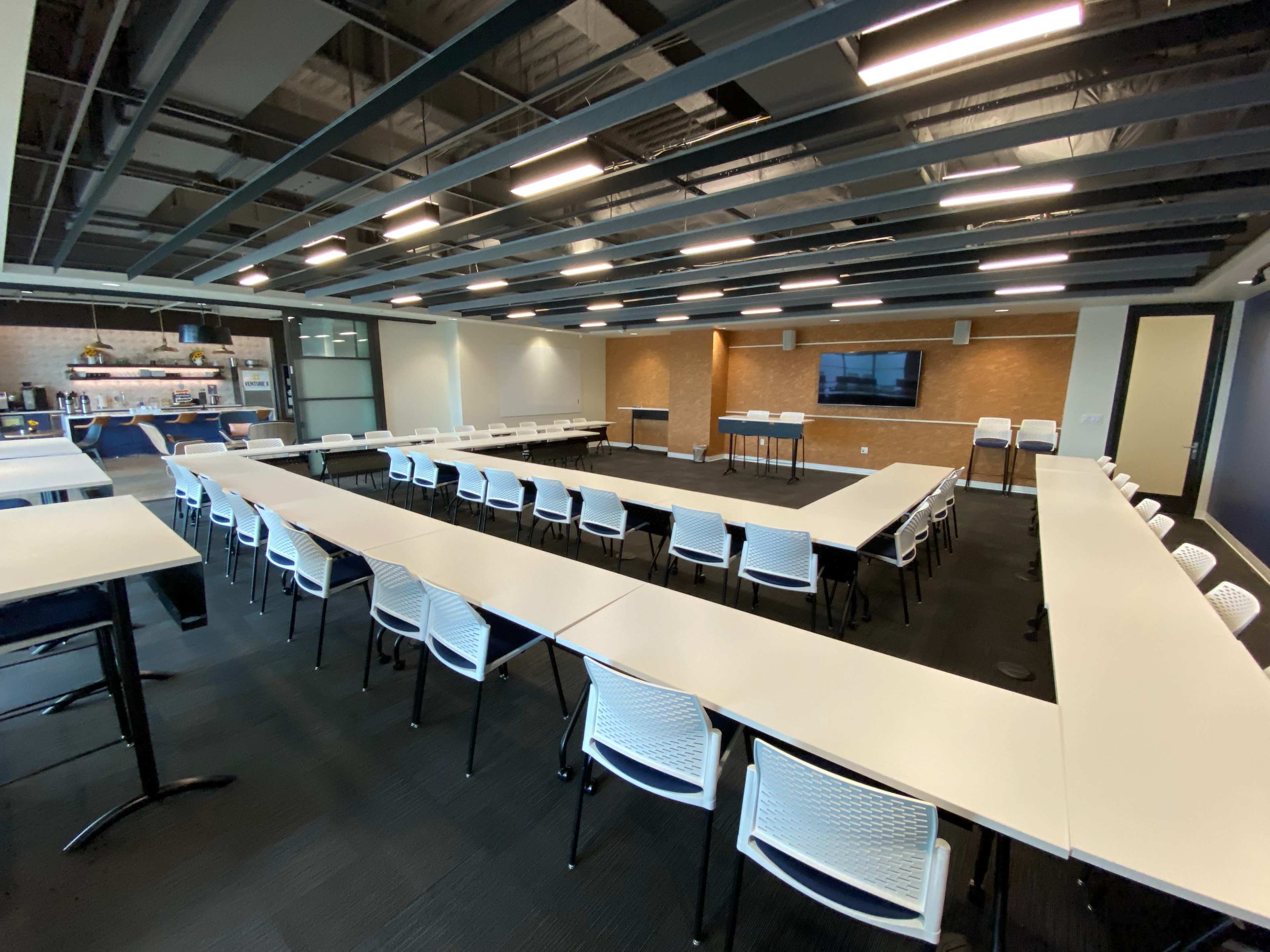 A modern conference room with multiple long tables arranged in a U-shape, facing a screen on the wall.