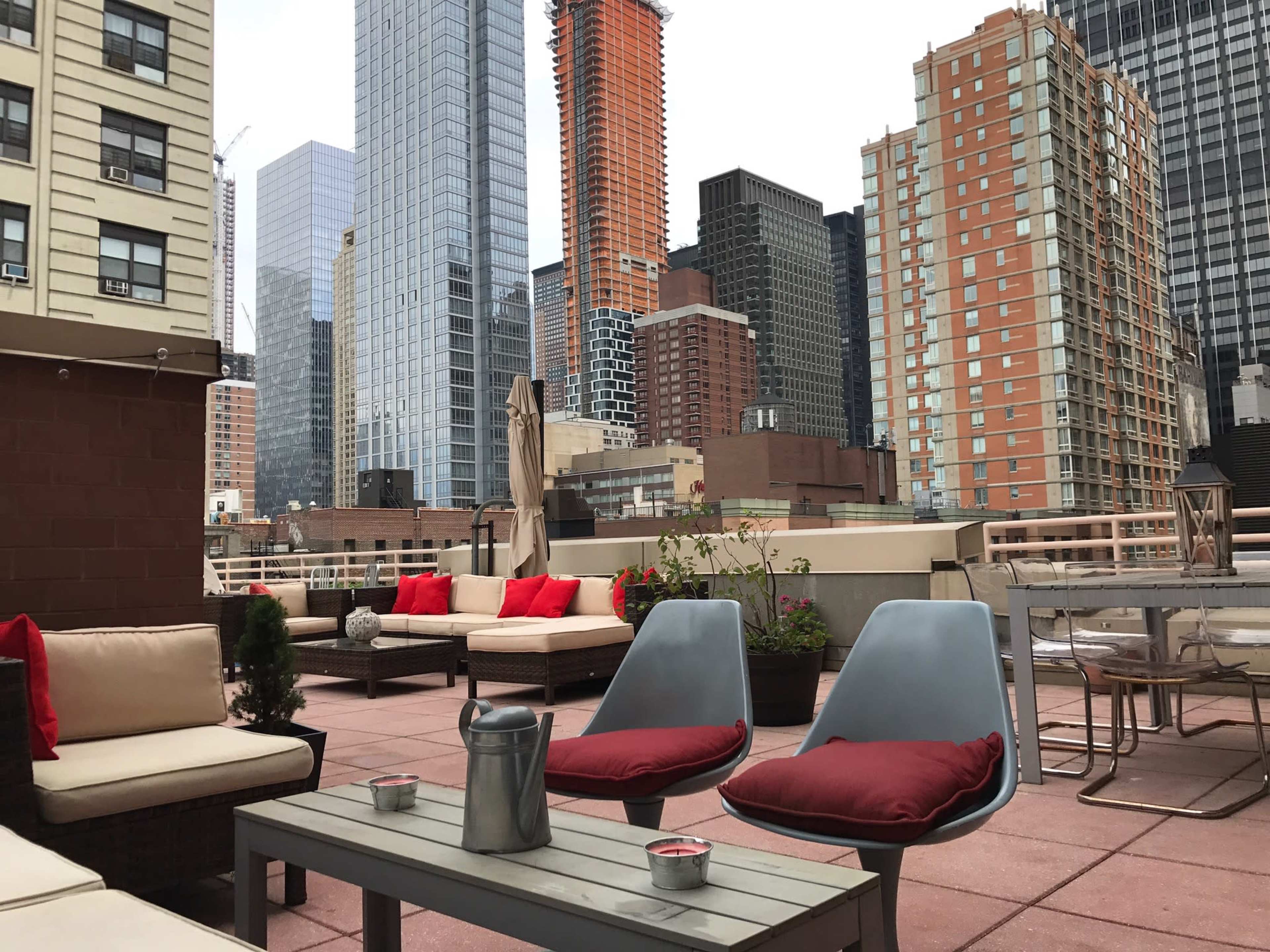 A rooftop terrace features seating arrangements with cushions, landscaped planters, and a skyline of tall buildings in the background.