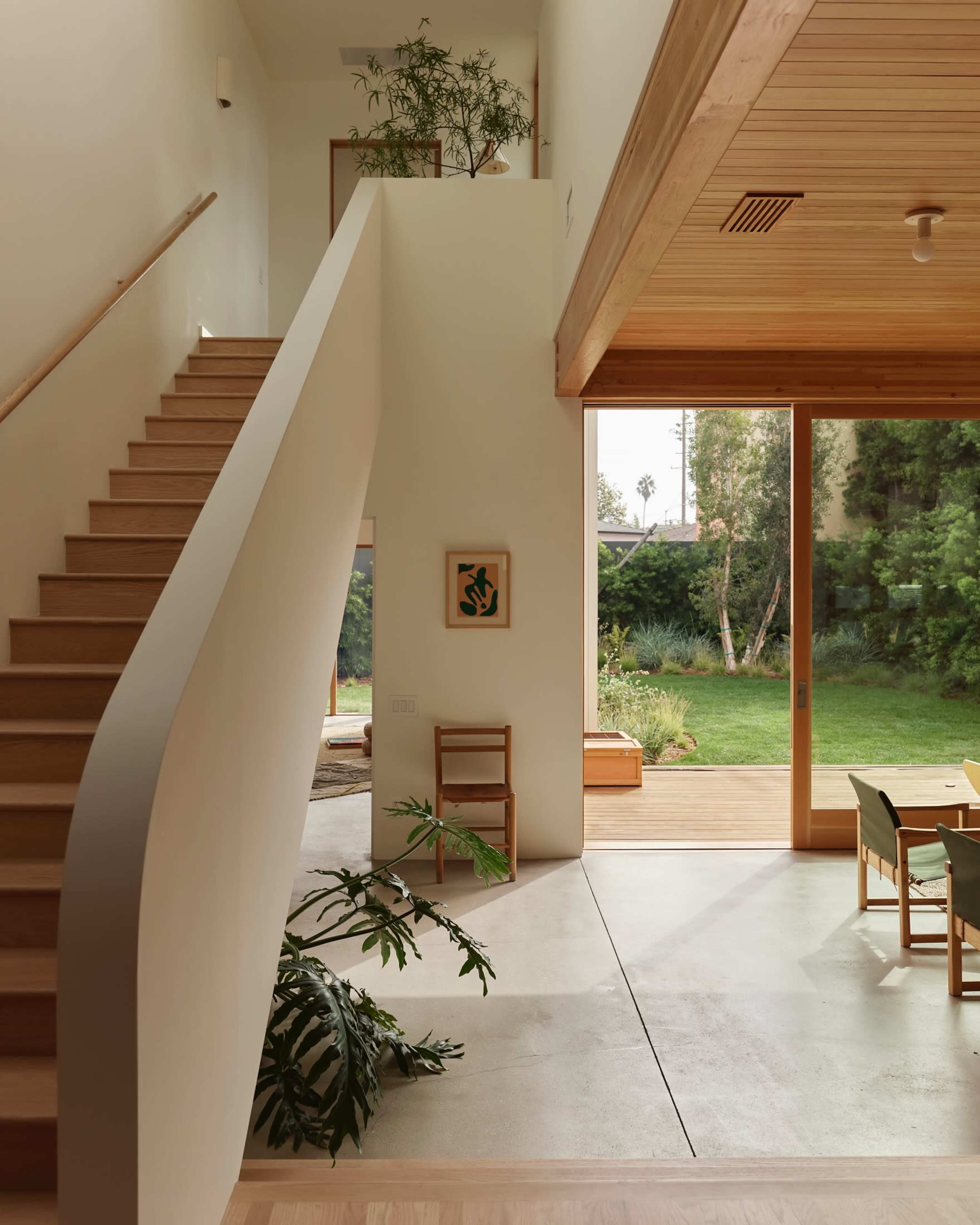 A bright interior features a staircase leading to an upper level, with large windows opening to a garden and modern furnishings.
