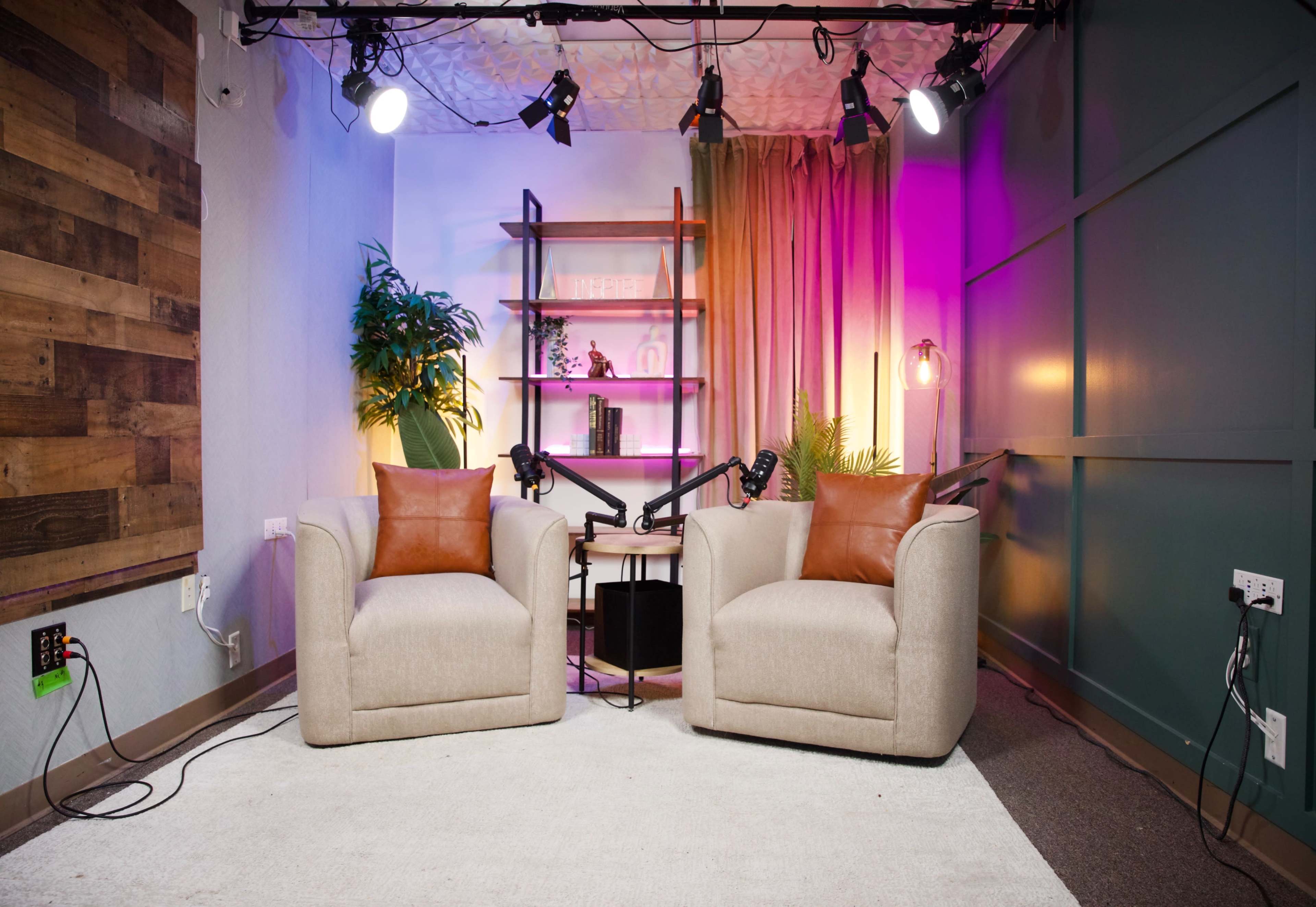 The image shows a cozy studio setup with two beige armchairs facing a small table, surrounded by decorative shelves and colorful lighting.