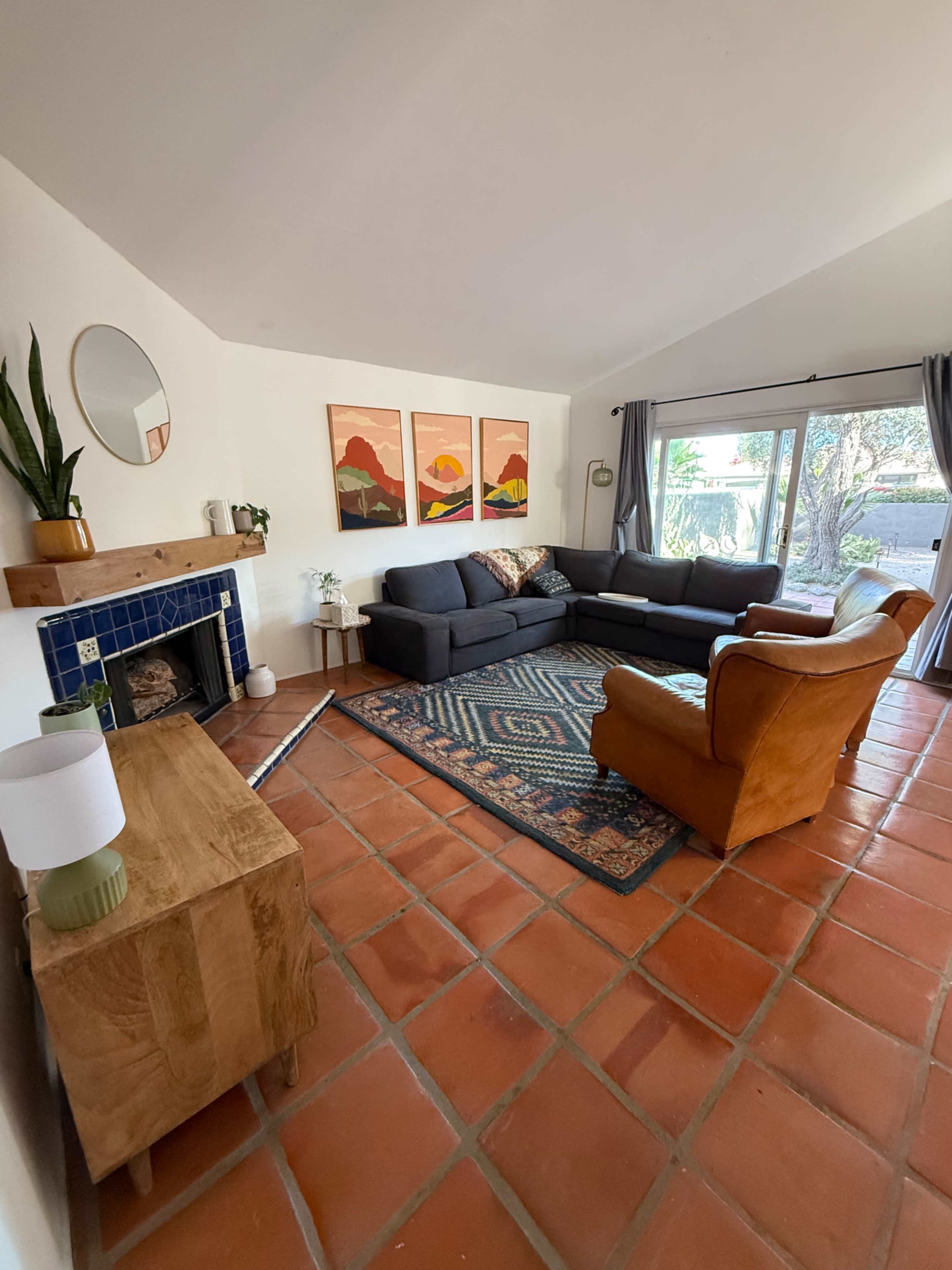The image shows a living room with a gray sectional sofa, a brown armchair, a wooden side table, and a fireplace, all arranged on a tiled floor with artwork on the walls and large windows allowing natural light in.
