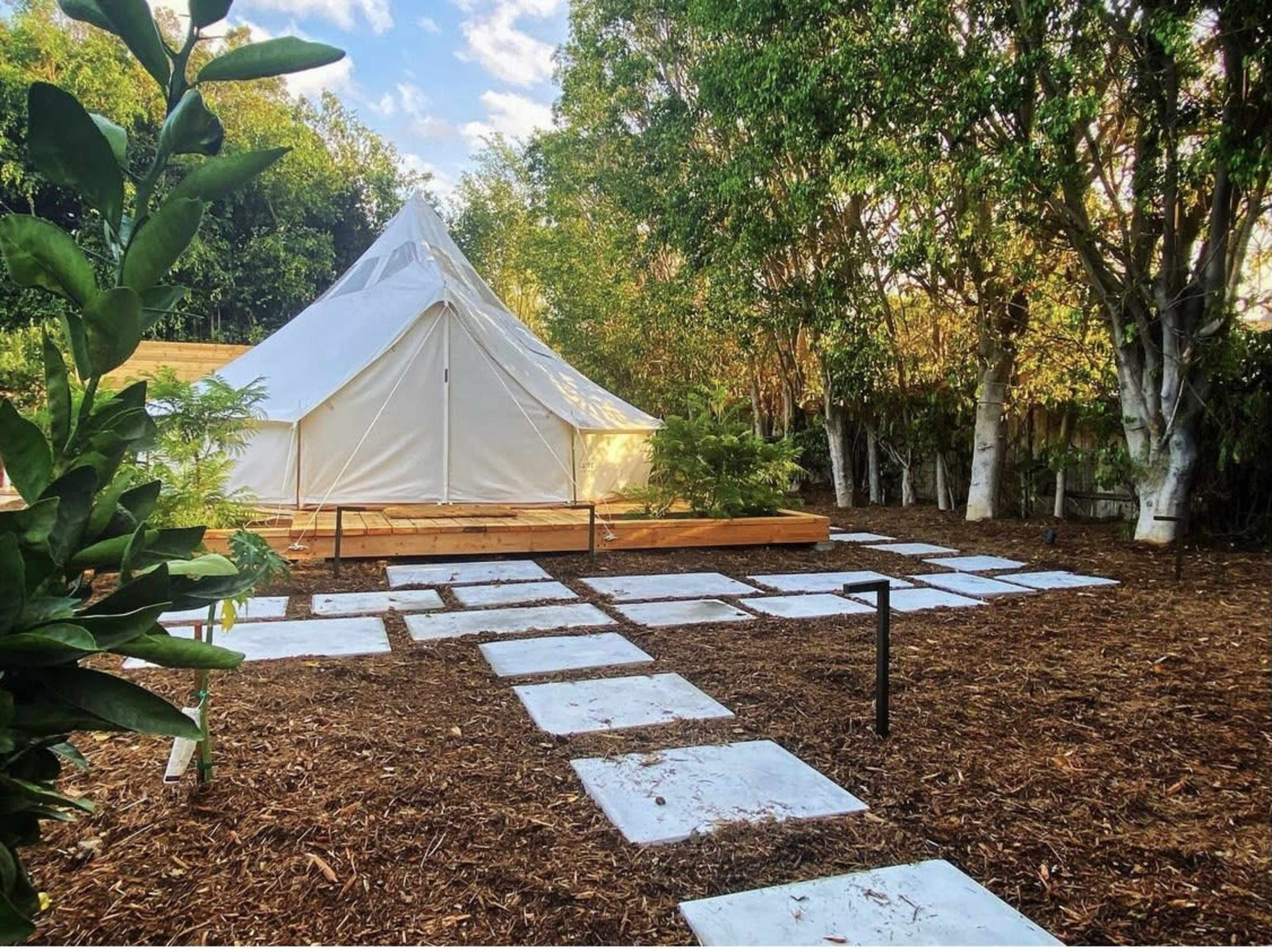 A white tent is set on a wooden platform surrounded by a pathway made of rectangular stones, amid greenery and trees.