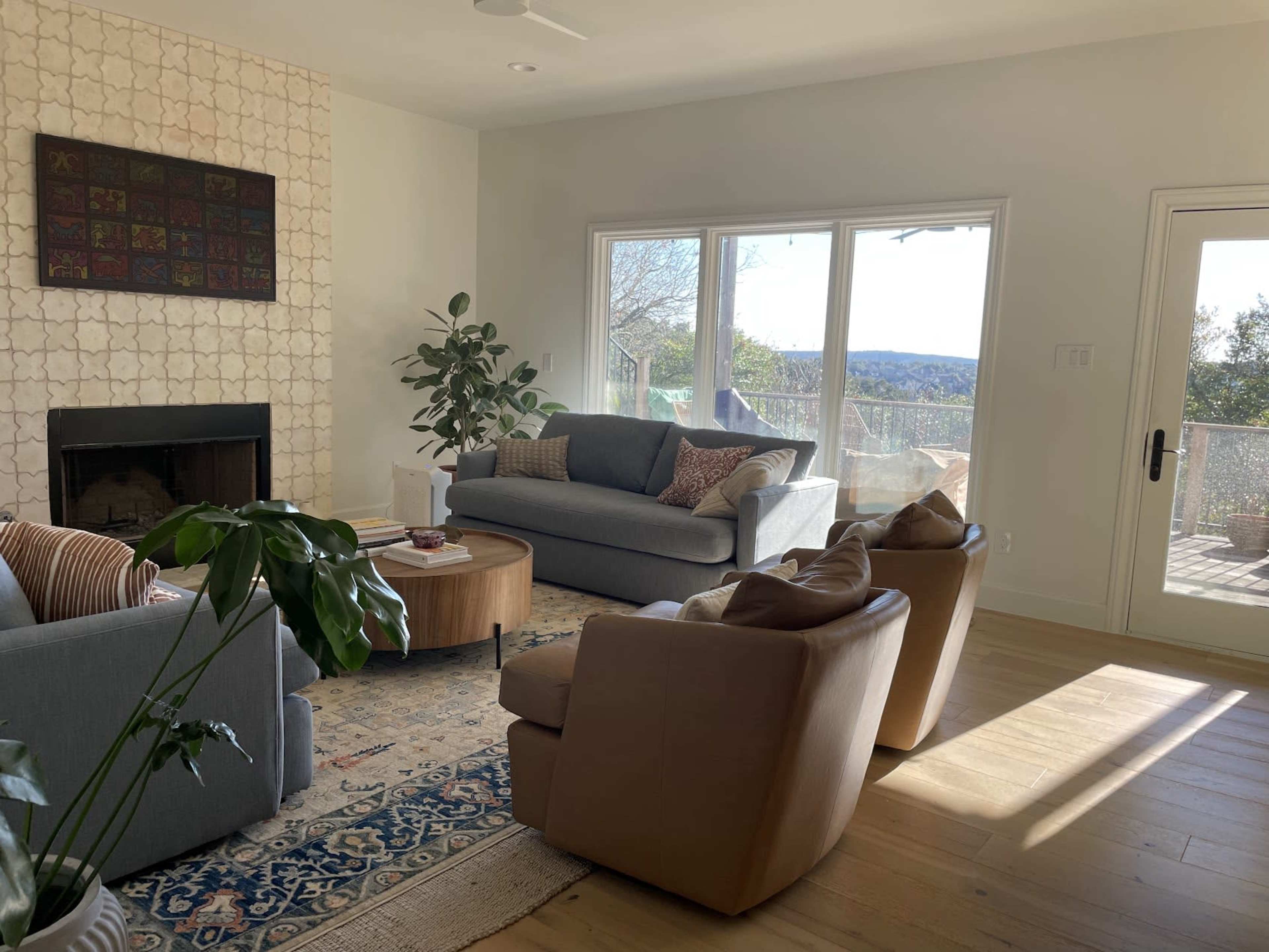 A spacious living room features a gray sofa and two tan armchairs surrounding a round coffee table, with large windows providing a view of the outdoors.
