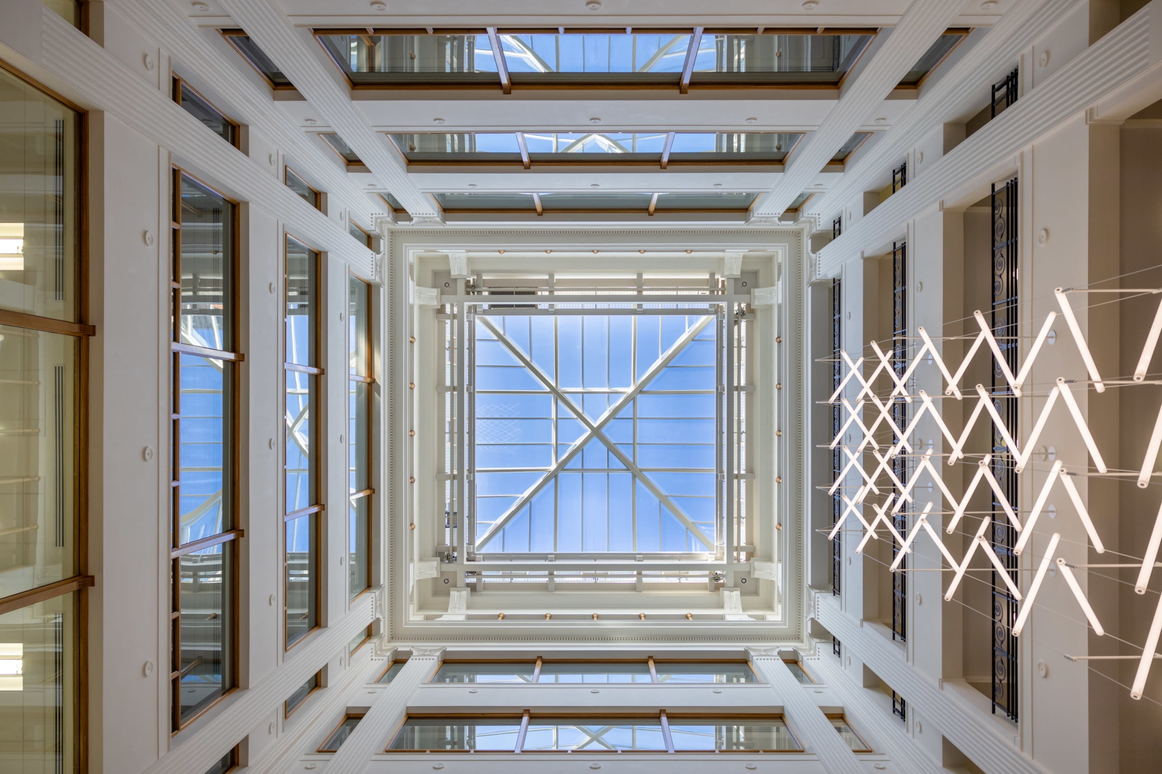 An interior atrium with a glass ceiling and linear lighting, viewed from below.