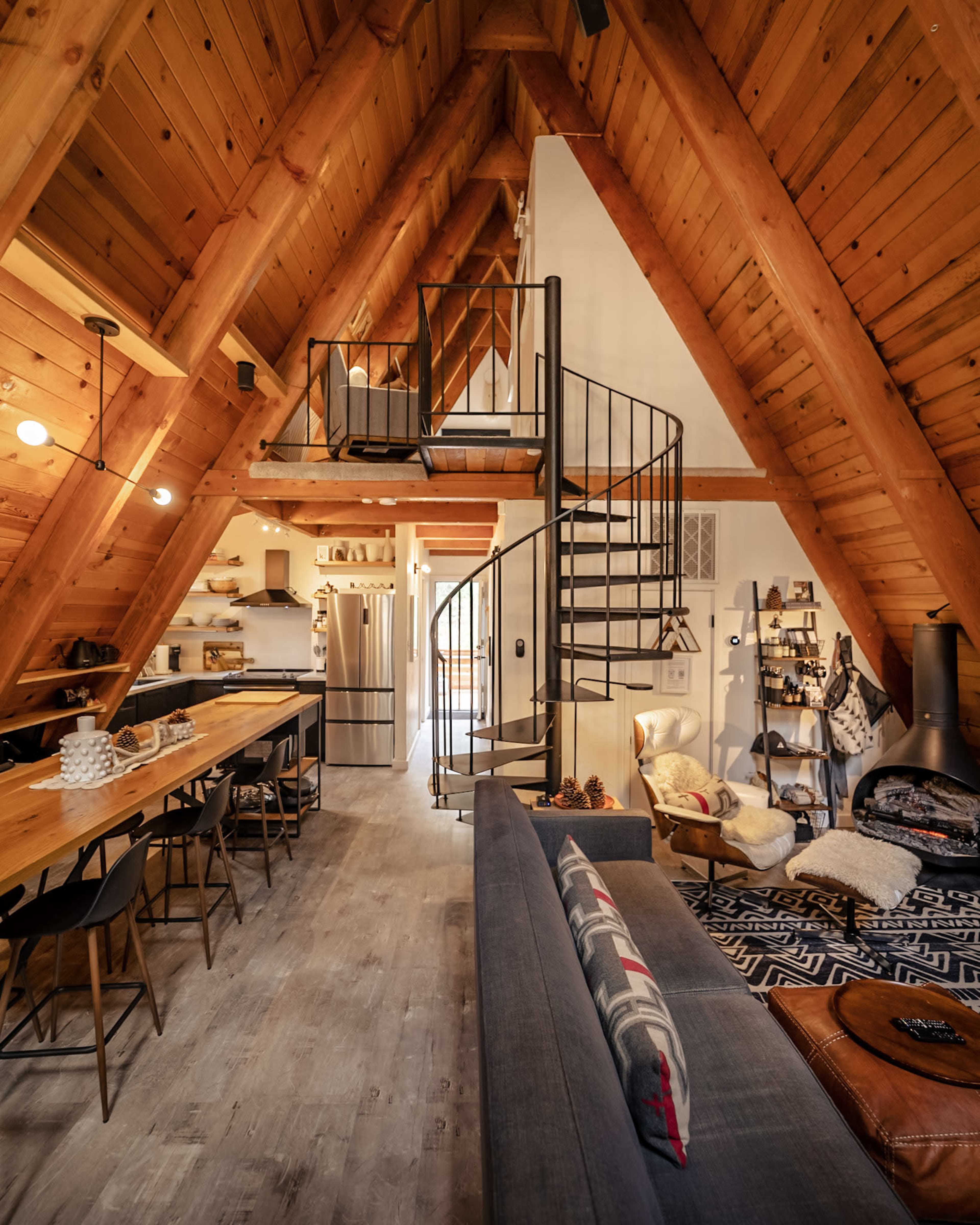 The image shows an interior of a modern A-frame cabin featuring a spiral staircase, wooden beams, a dining table, and a cozy living area.