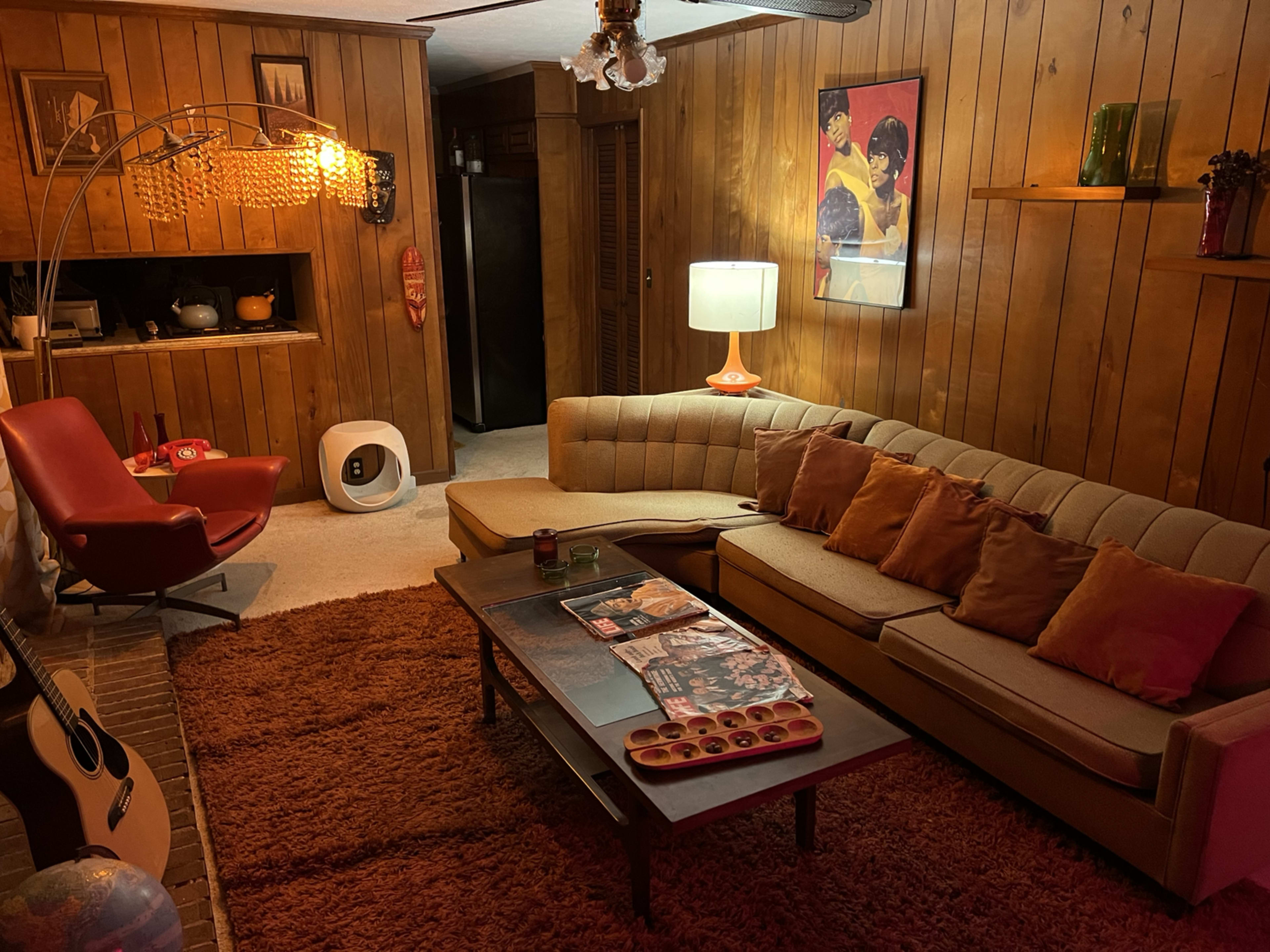 The image shows a cozy living room with wooden paneling, a sectional couch adorned with orange pillows, a floor lamp, and a coffee table featuring magazines and games.