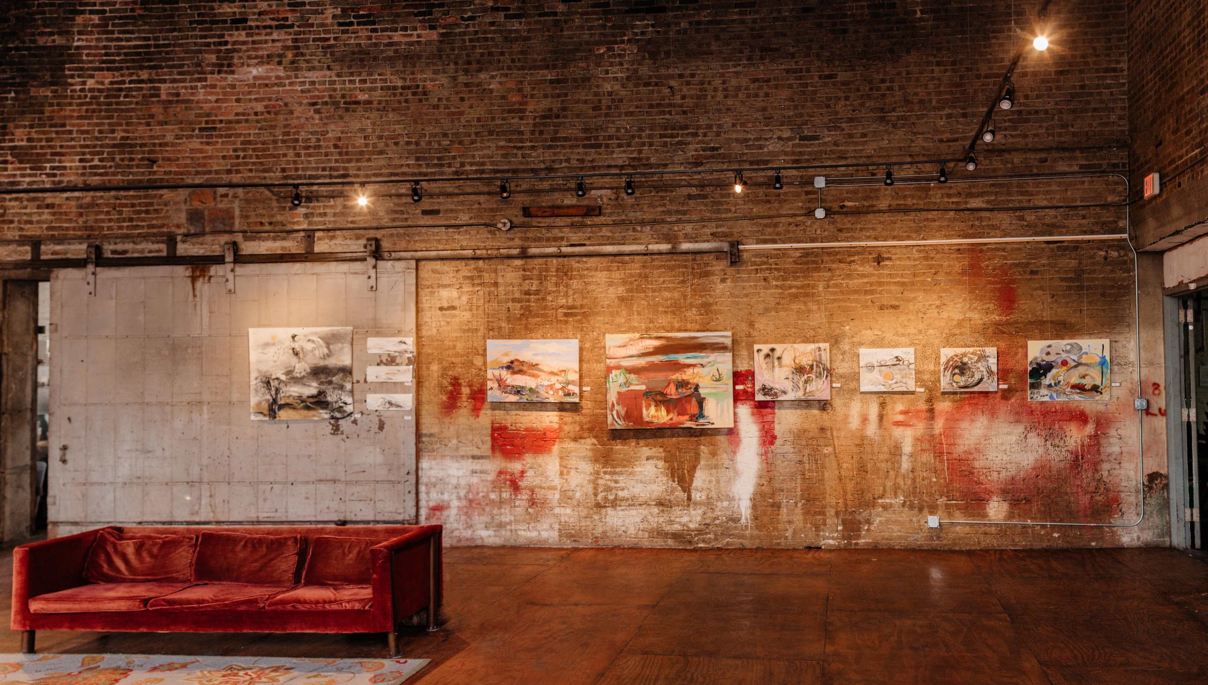 A gallery interior with a brick wall displaying several abstract paintings and a red velvet couch in the foreground.