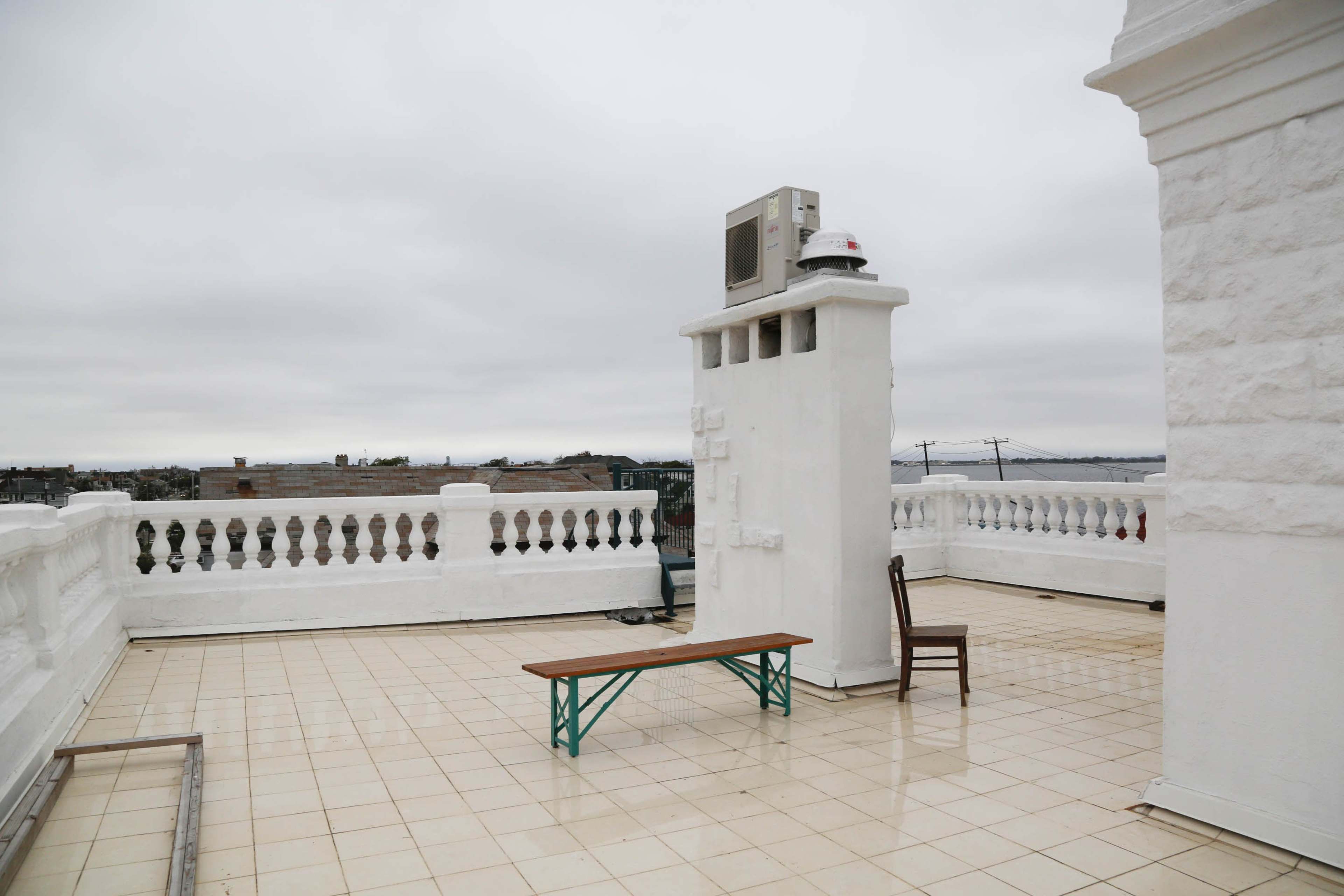 A rooftop terrace features a tiled floor with a wooden bench and a single chair, surrounded by white walls and an air conditioning unit.
