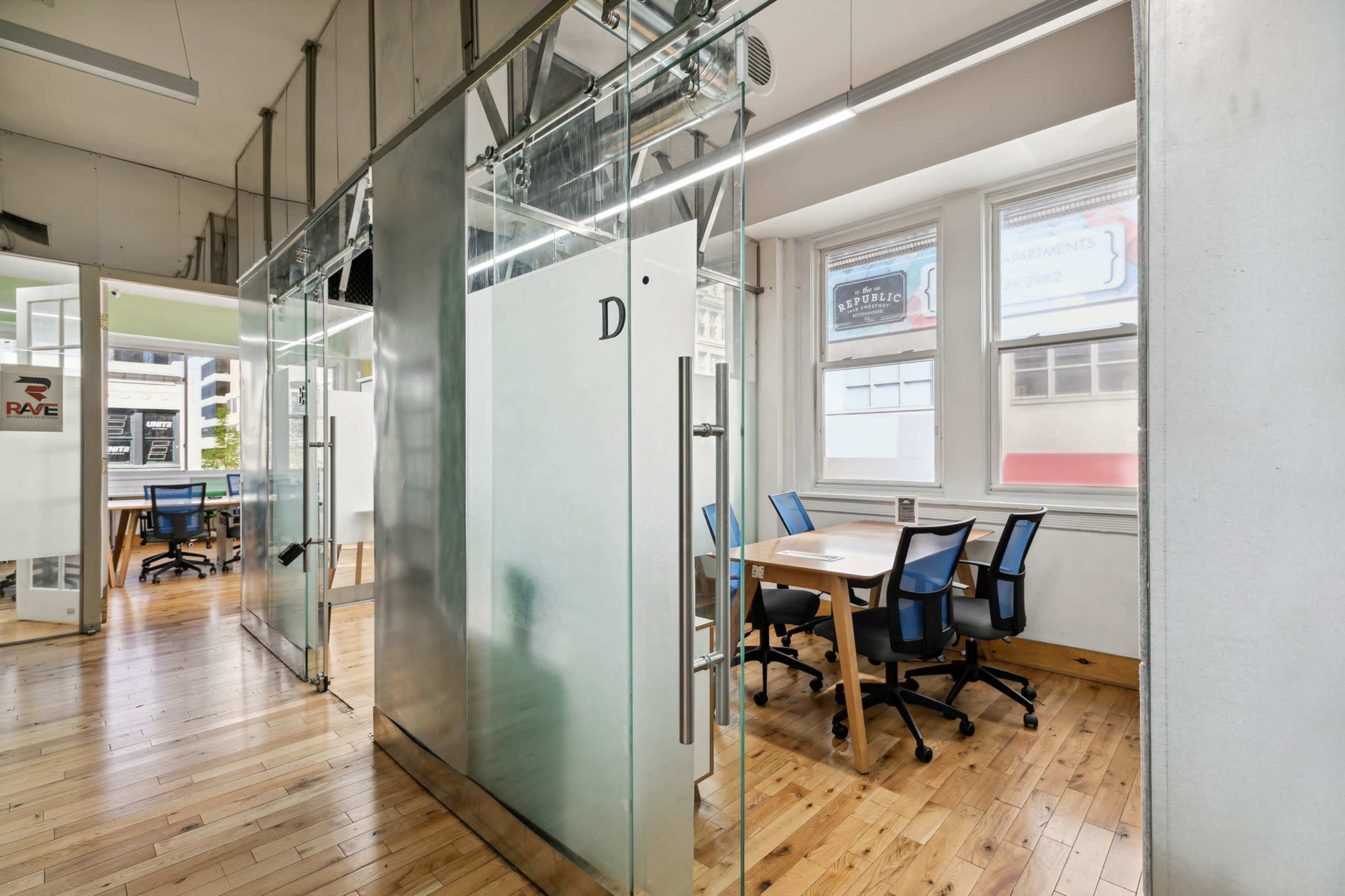 The image shows a modern office space with glass partitions, wooden flooring, and workstations arranged against the walls.