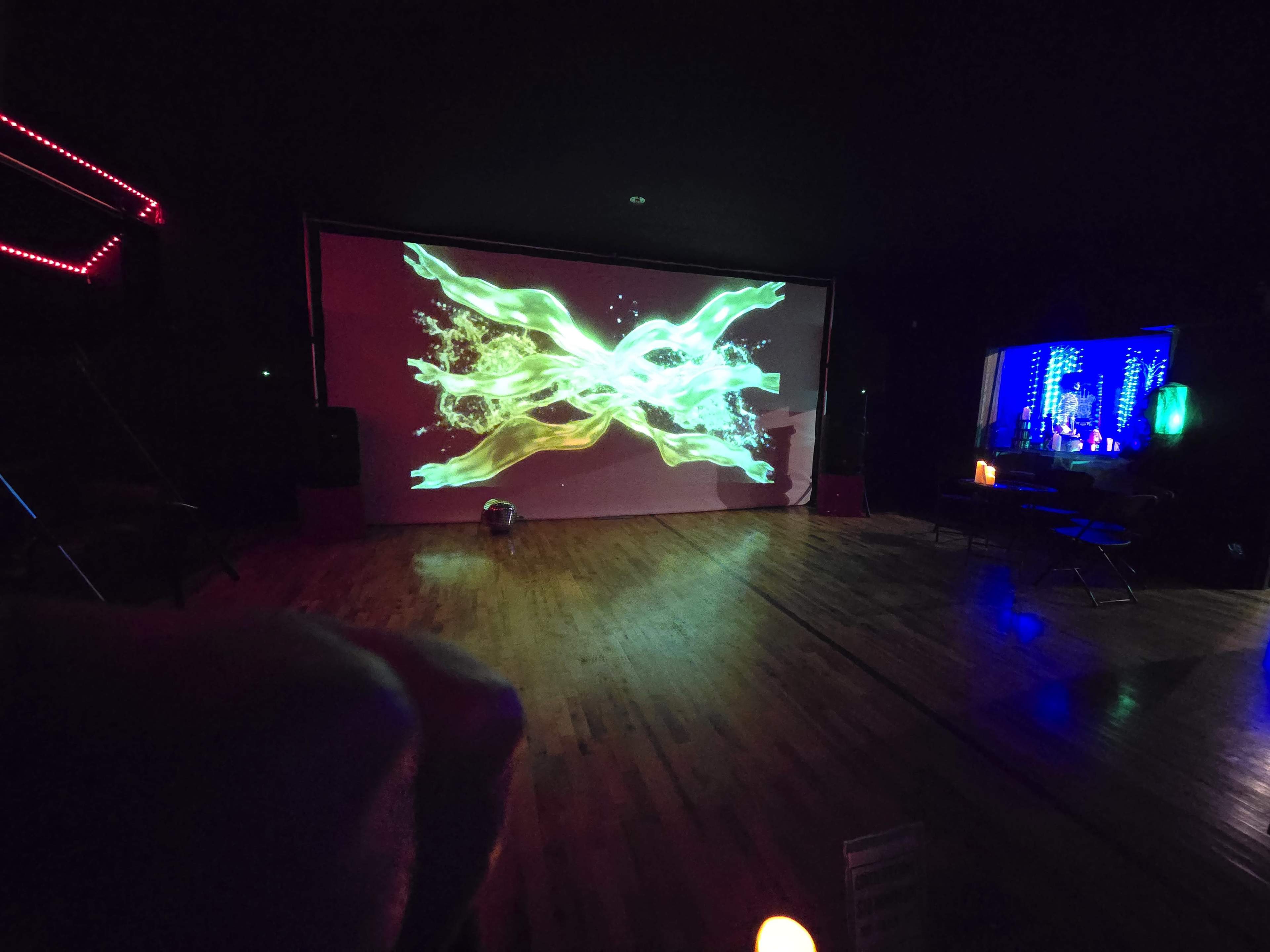 The room features a large digital projection of swirling green and yellow designs on a wall, with ambient lighting and a small table in the corner.