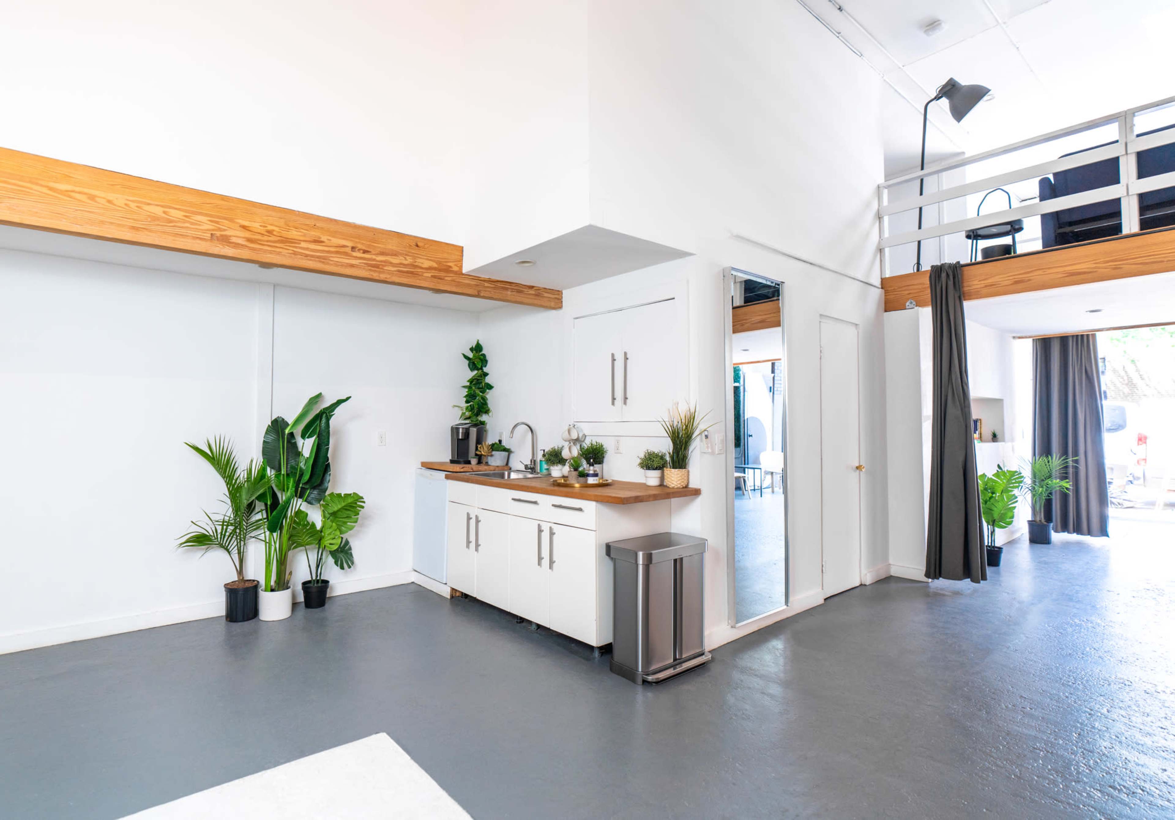 A modern, open-concept kitchen area with white cabinets, a wooden countertop, and several potted plants, leading to a seating area with a staircase in the background.