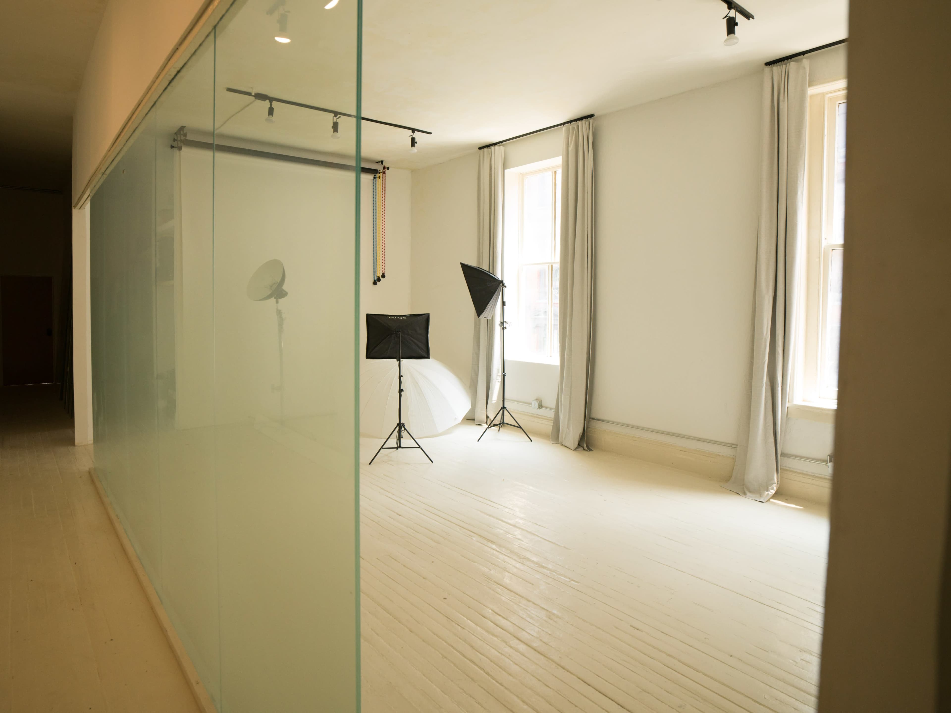 The image shows a bright photo studio with white walls, natural light coming through large windows, and studio lighting equipment set up on the floor.