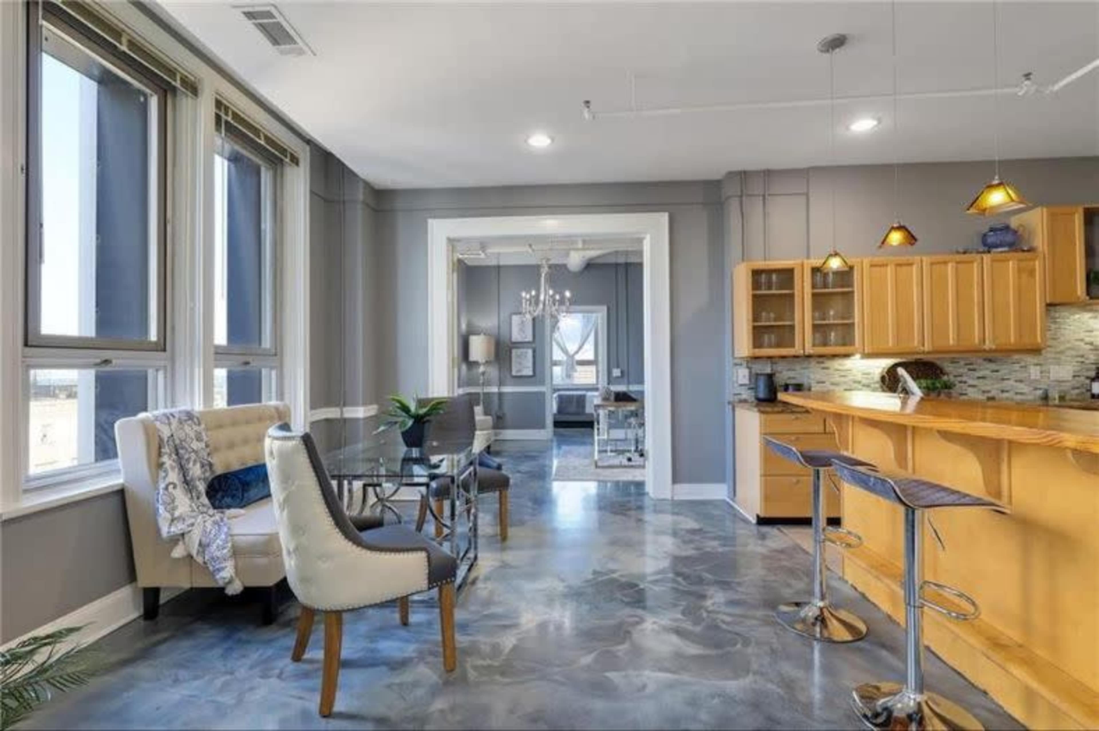 The image shows a modern, open-concept living area with a kitchen bar, seating arrangements, and large windows allowing natural light to fill the space.
