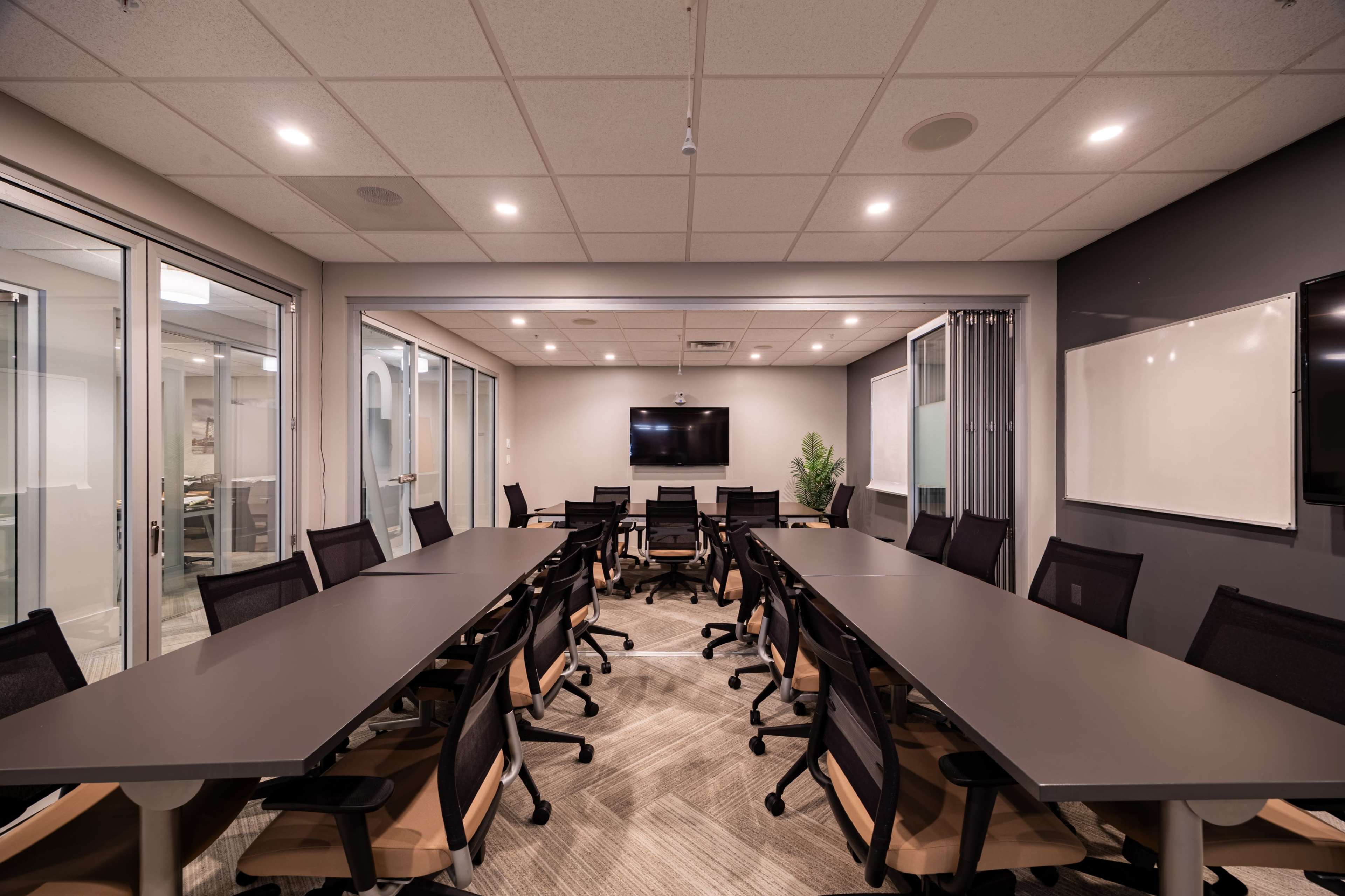 The image shows a modern conference room with a long table, multiple chairs, and audio-visual equipment mounted on the walls.