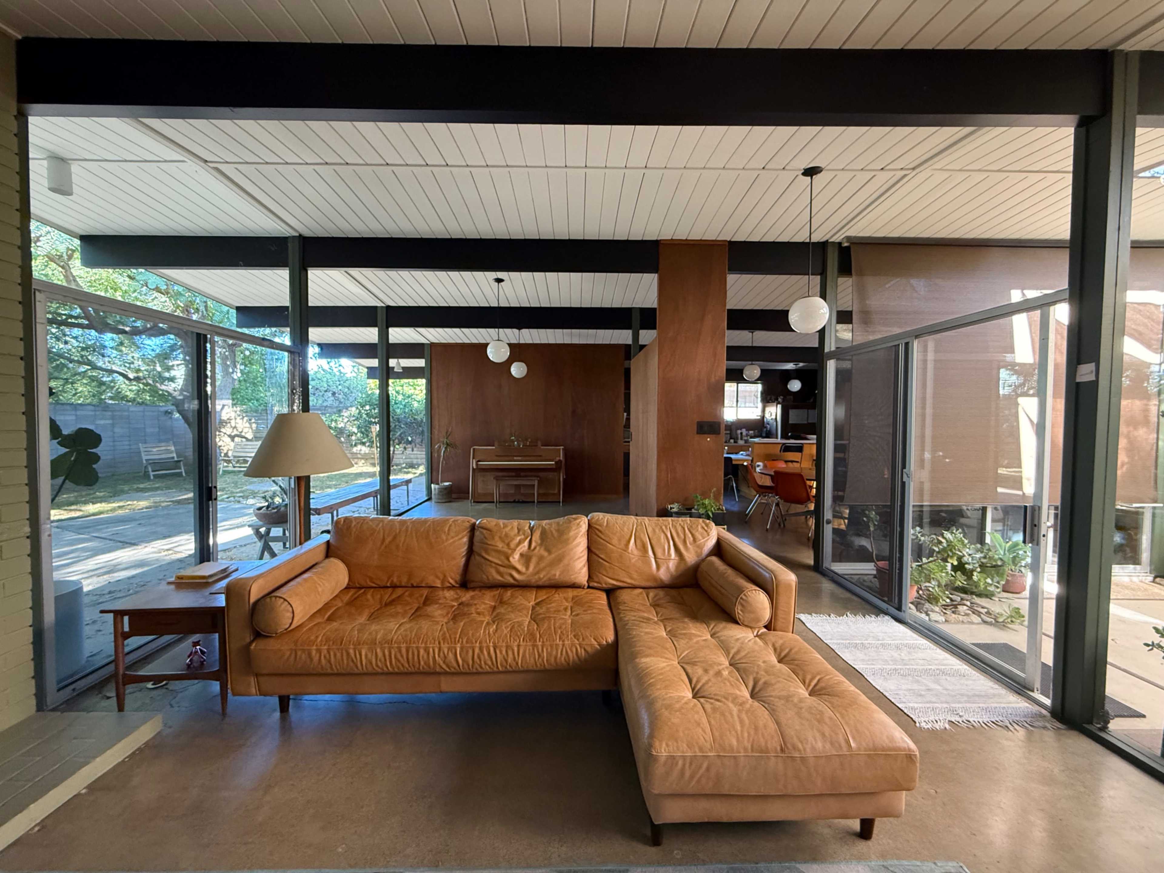 The image shows a modern living room featuring a large brown leather sectional sofa, large glass windows, and a view of an outdoor garden.