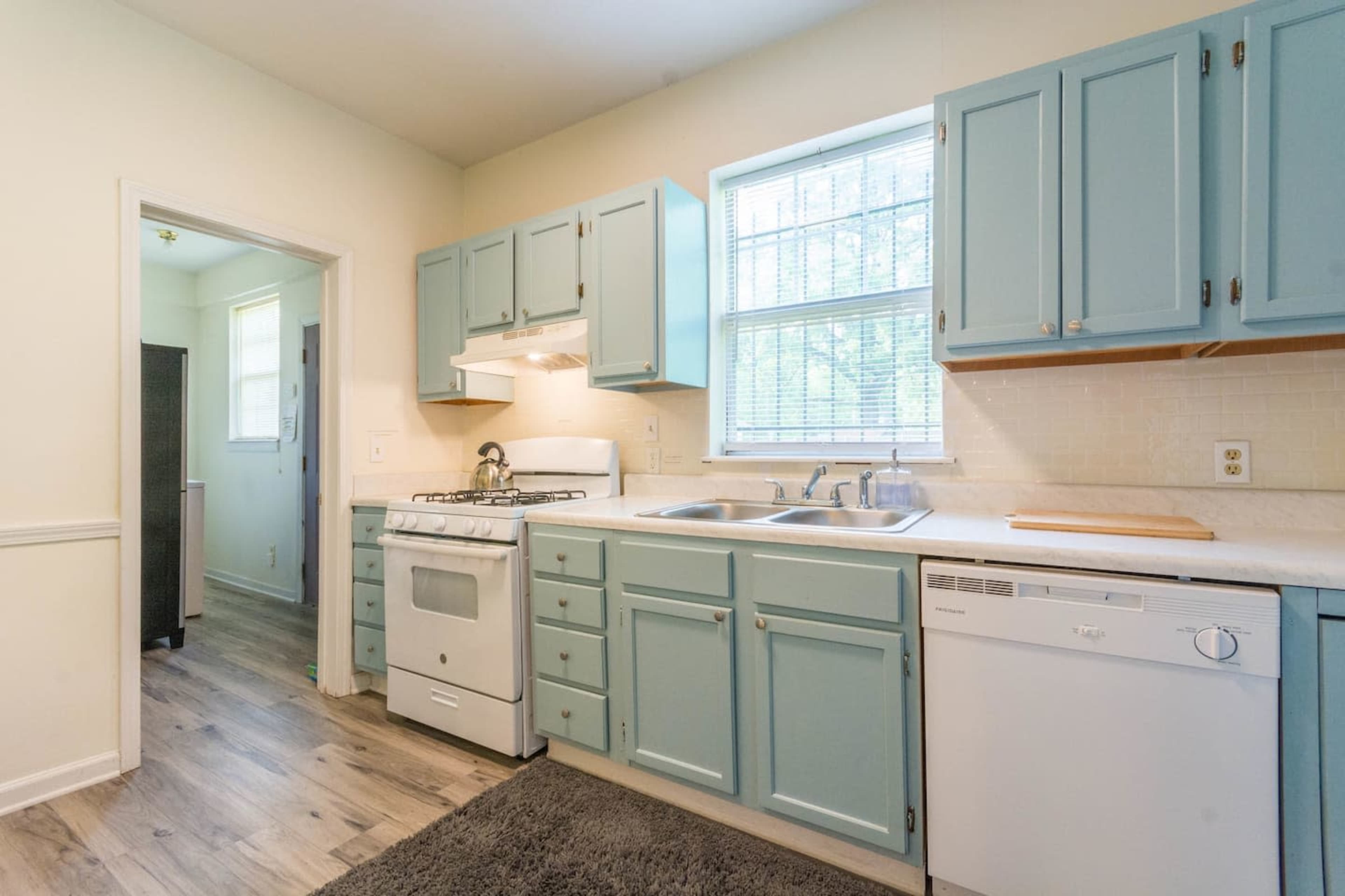 The kitchen features light blue cabinetry, a white countertop, and stainless steel appliances, with a large window above the sink providing natural light.