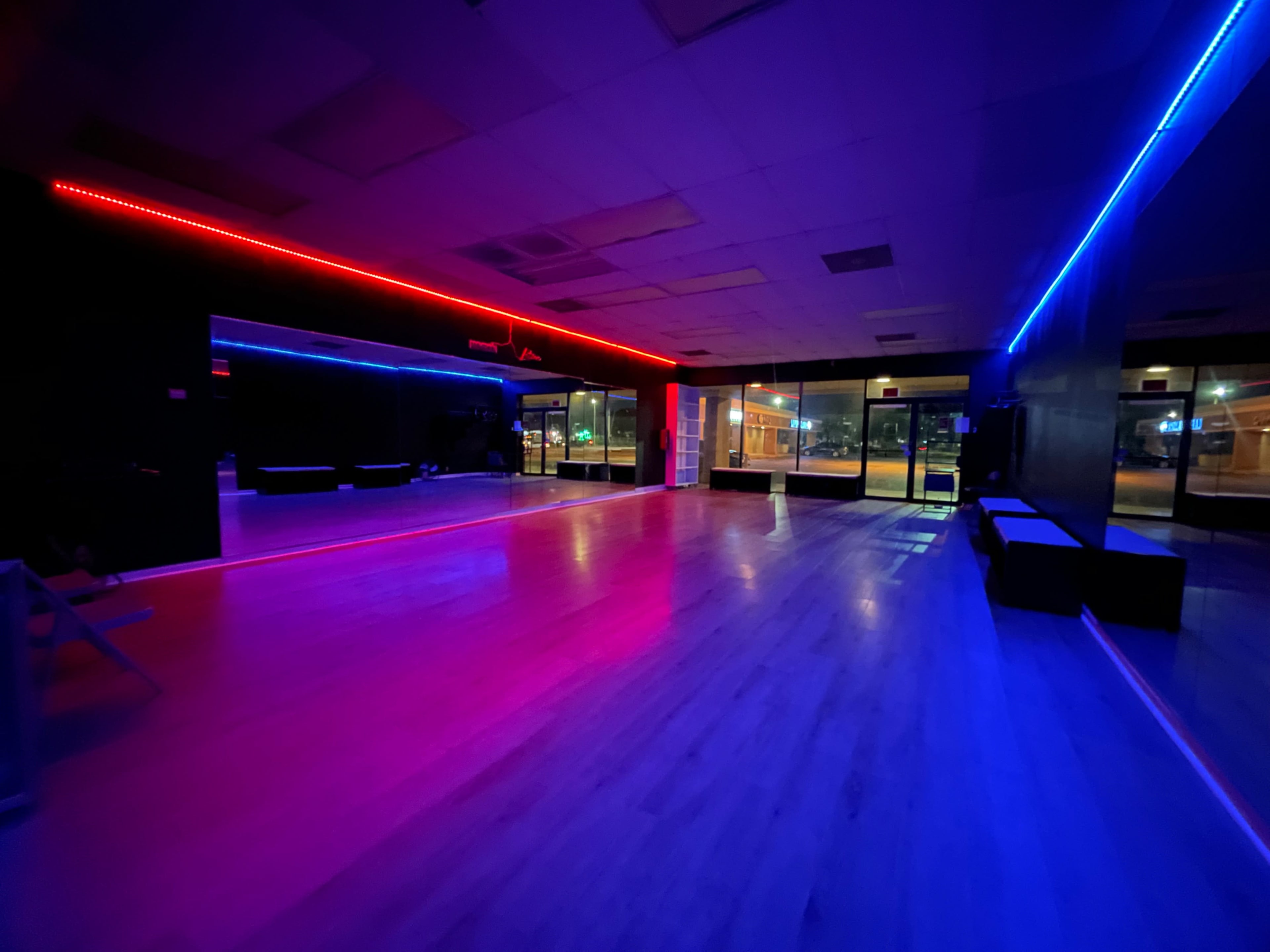 A dimly lit indoor studio features colorful LED lights in red and blue along the walls, with a polished wooden floor and large windows facing an outdoor area.