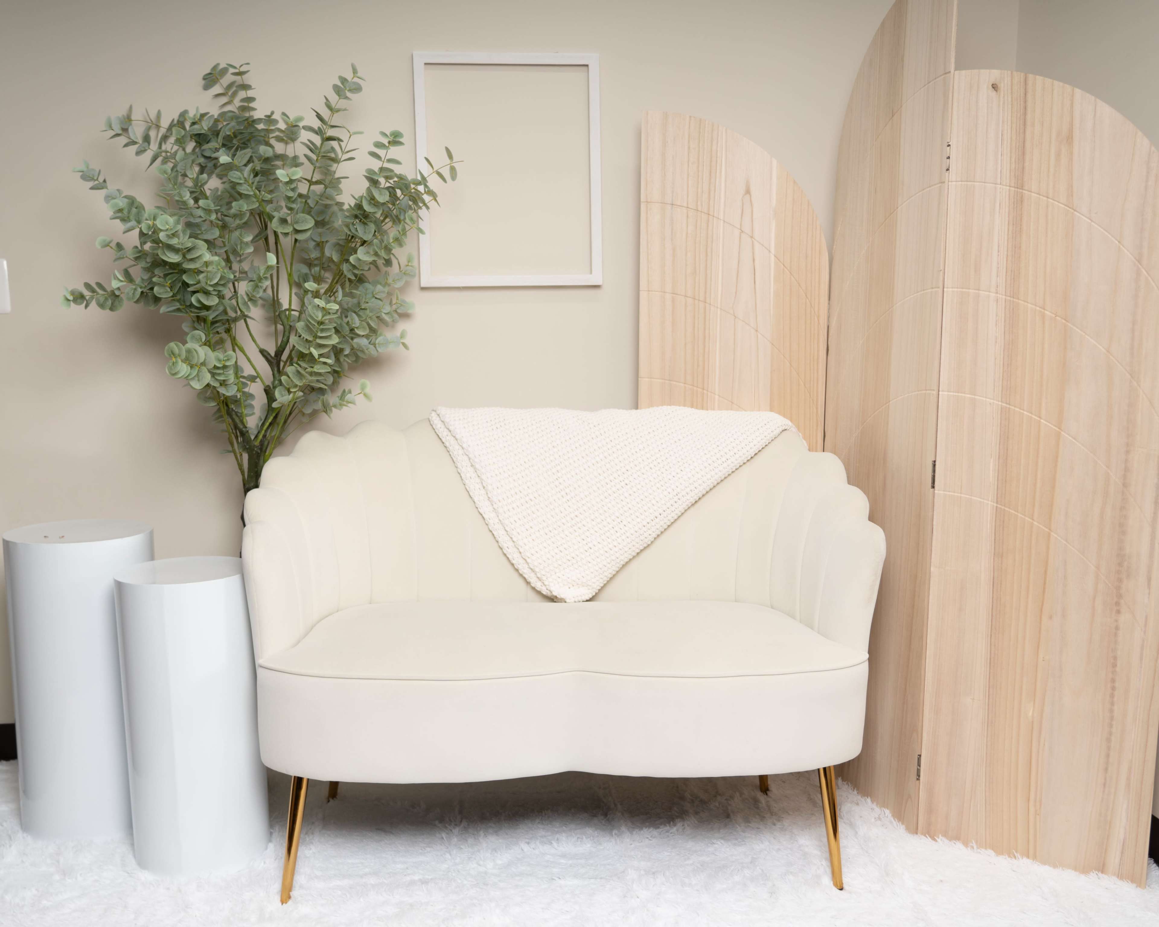 A cream-colored loveseat with a blanket draped over it is placed beside a leafy plant and wooden room dividers in a minimalist setting.