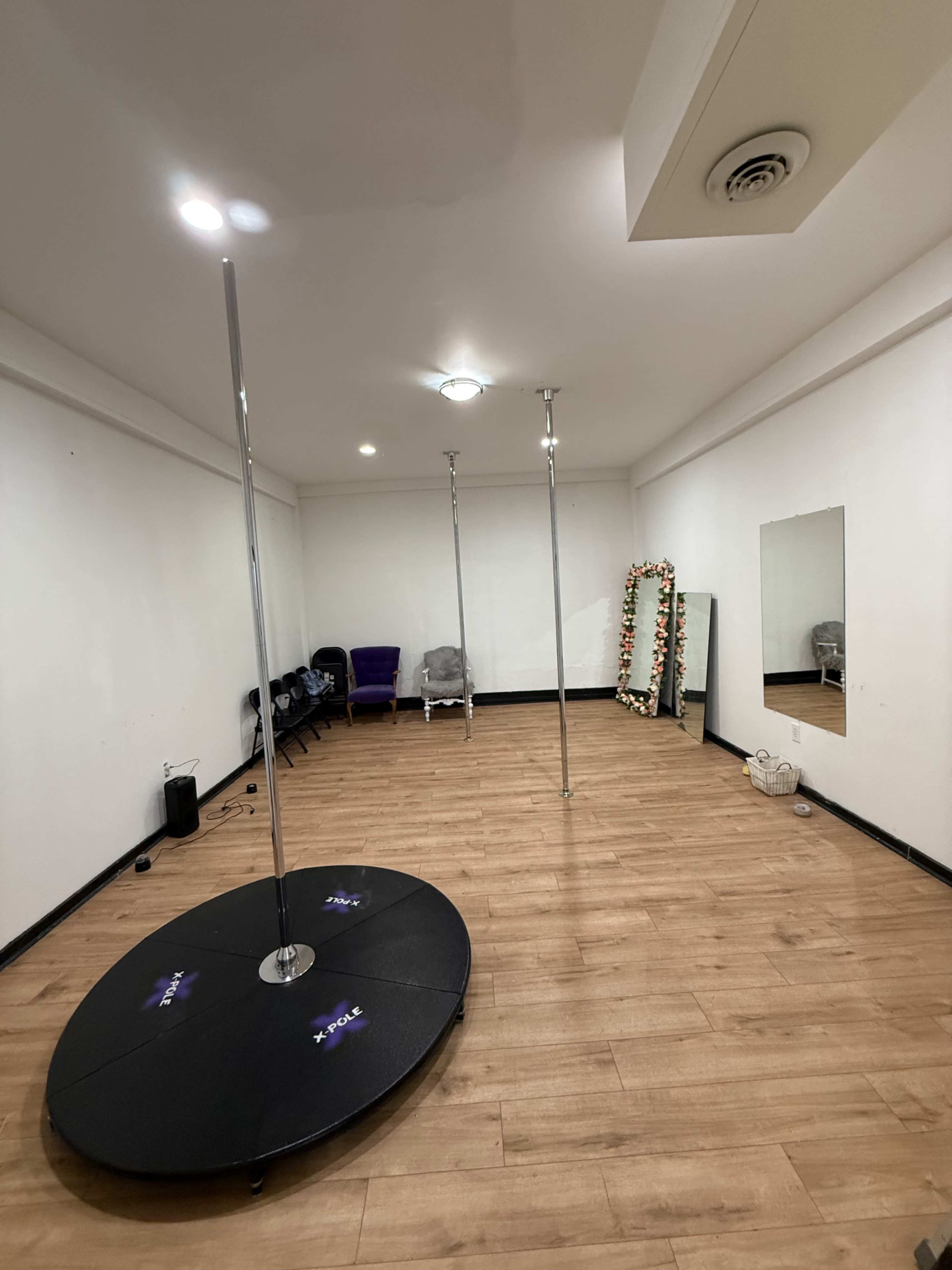 The image shows a spacious, empty studio with three pole dance poles, a mirror, and wooden flooring.