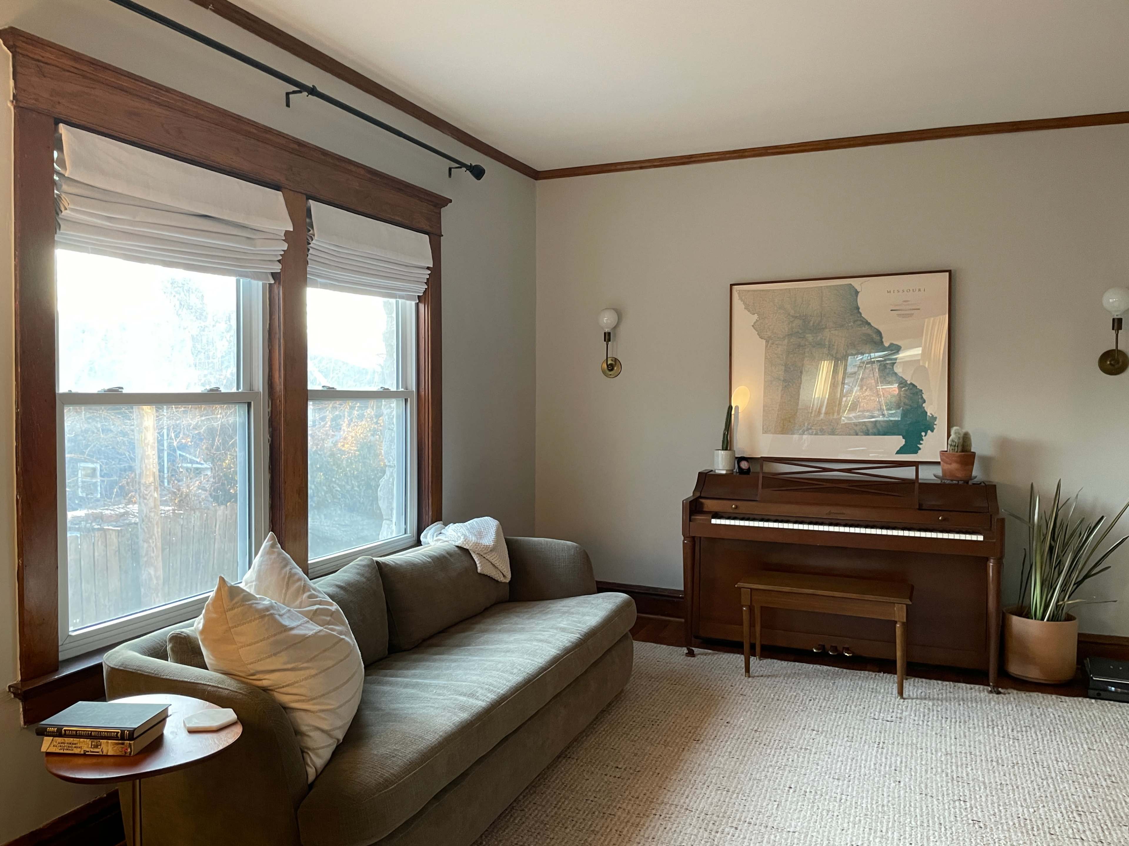 The image shows a cozy living room with a sofa, a piano, and large windows letting in natural light.