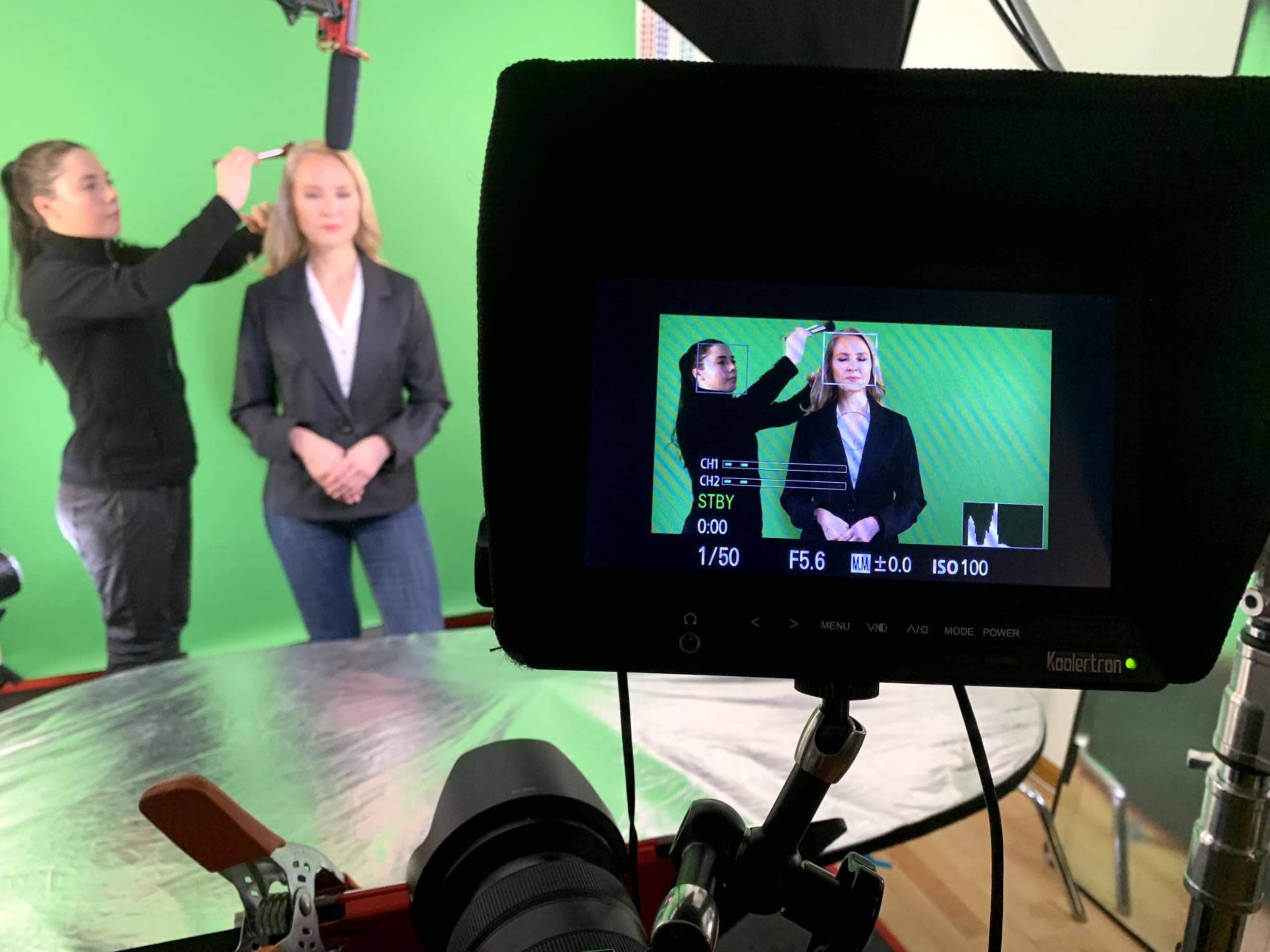 A woman is getting her makeup done in front of a green screen while another woman adjusts a camera setup.