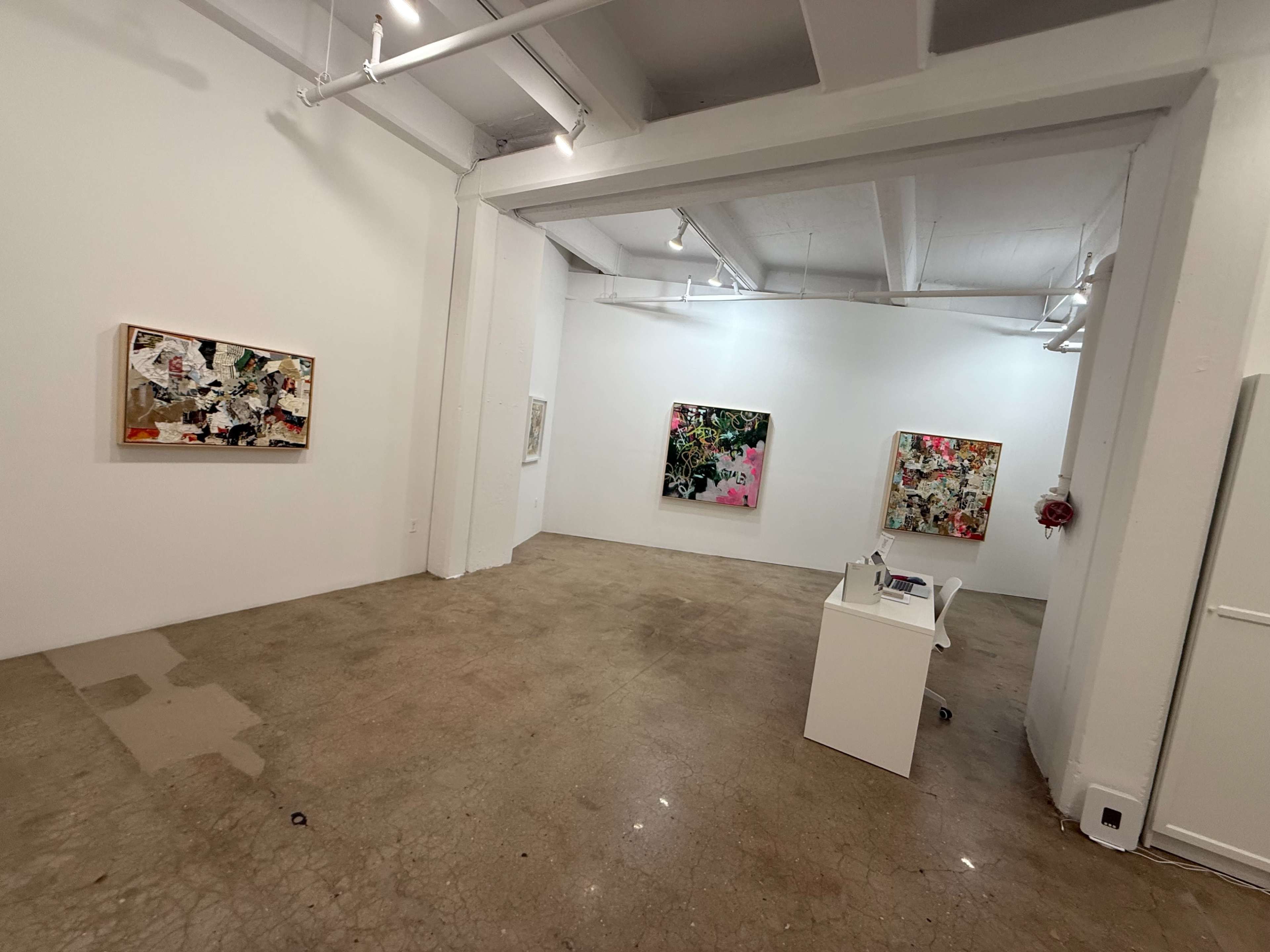 The image shows a spacious art gallery featuring several colorful abstract paintings on white walls, with a small desk positioned in the foreground.