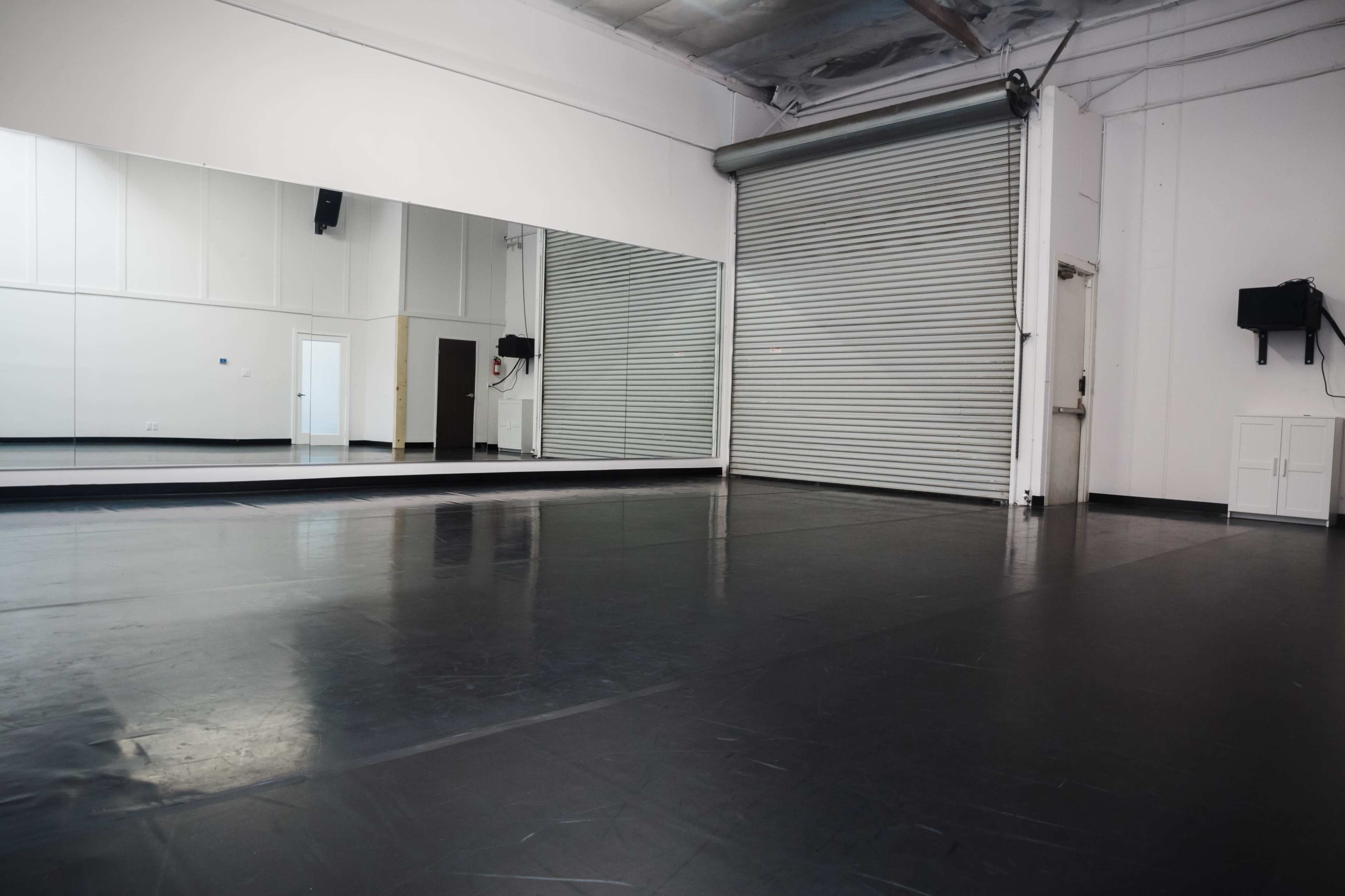 A spacious dance studio featuring a large mirror, a roll-up metal door, and a smooth black floor.