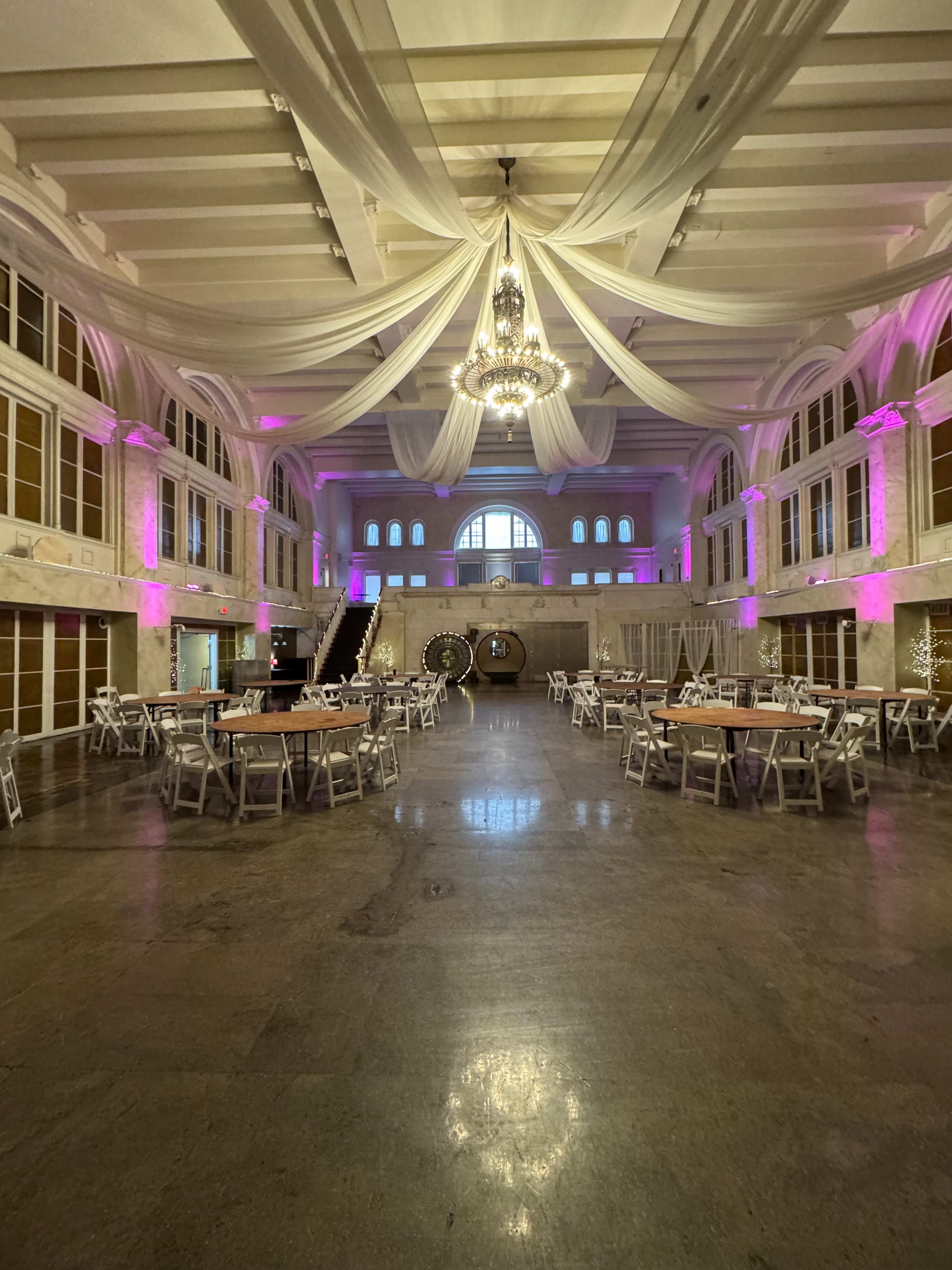 The image shows a spacious indoor event venue with decorative ceiling drapery, a chandelier, and tables arranged for a gathering.
