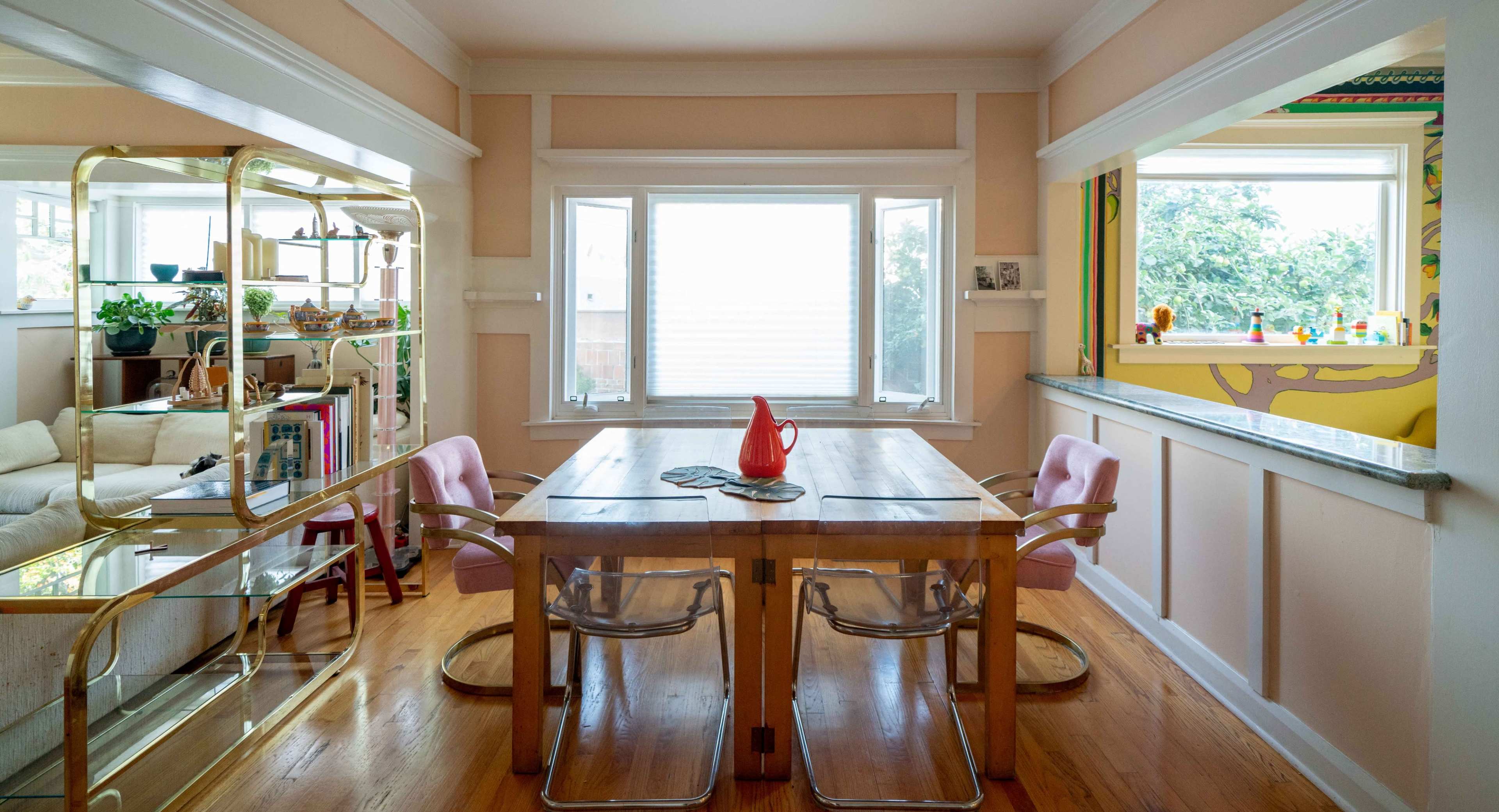 A dining area features a wooden table surrounded by clear chairs, with a large window and a shelf displaying plants and decorative items.