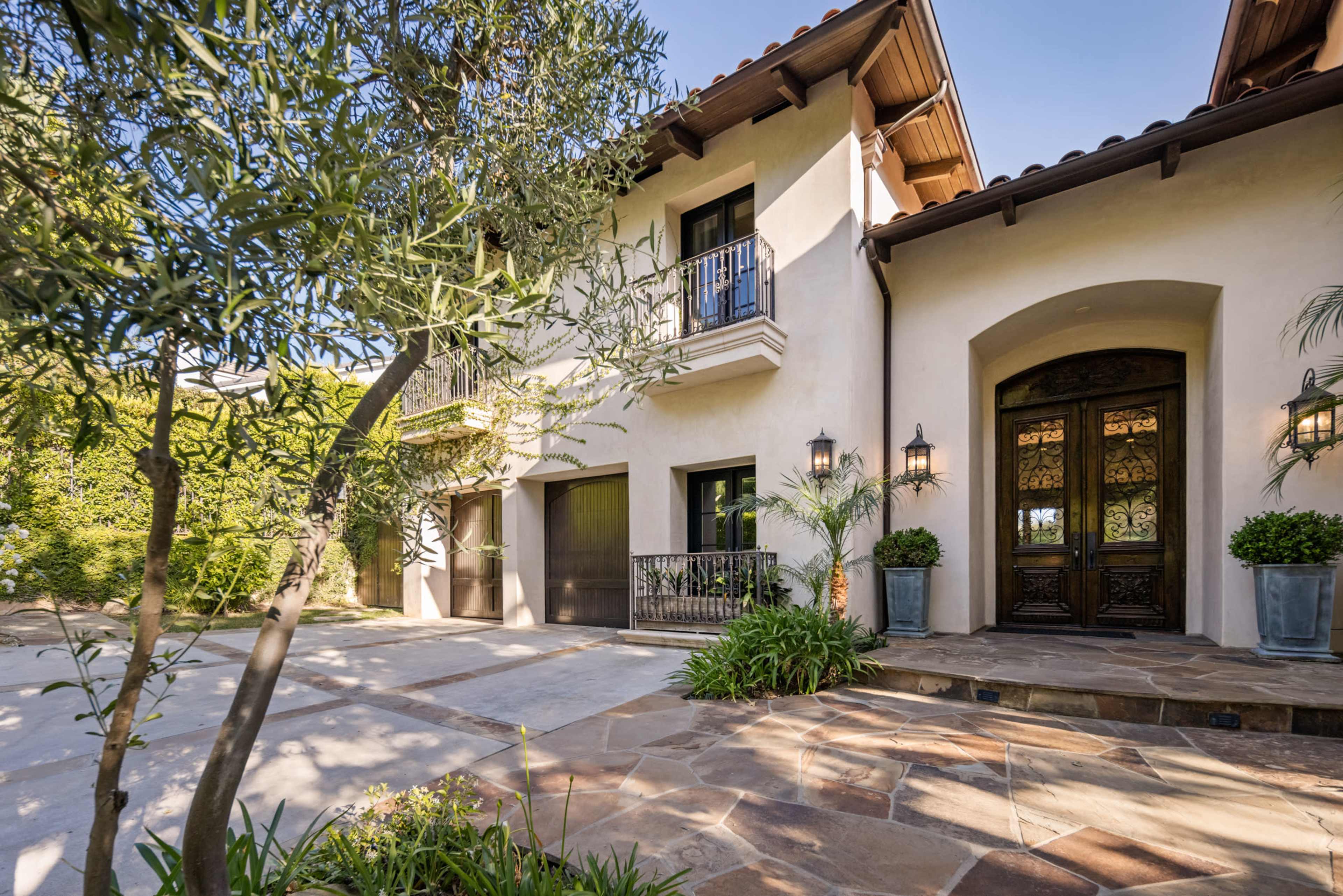 The image shows a two-story house with a stone pathway leading to a wooden front door, flanked by decorative plants and outdoor lighting.