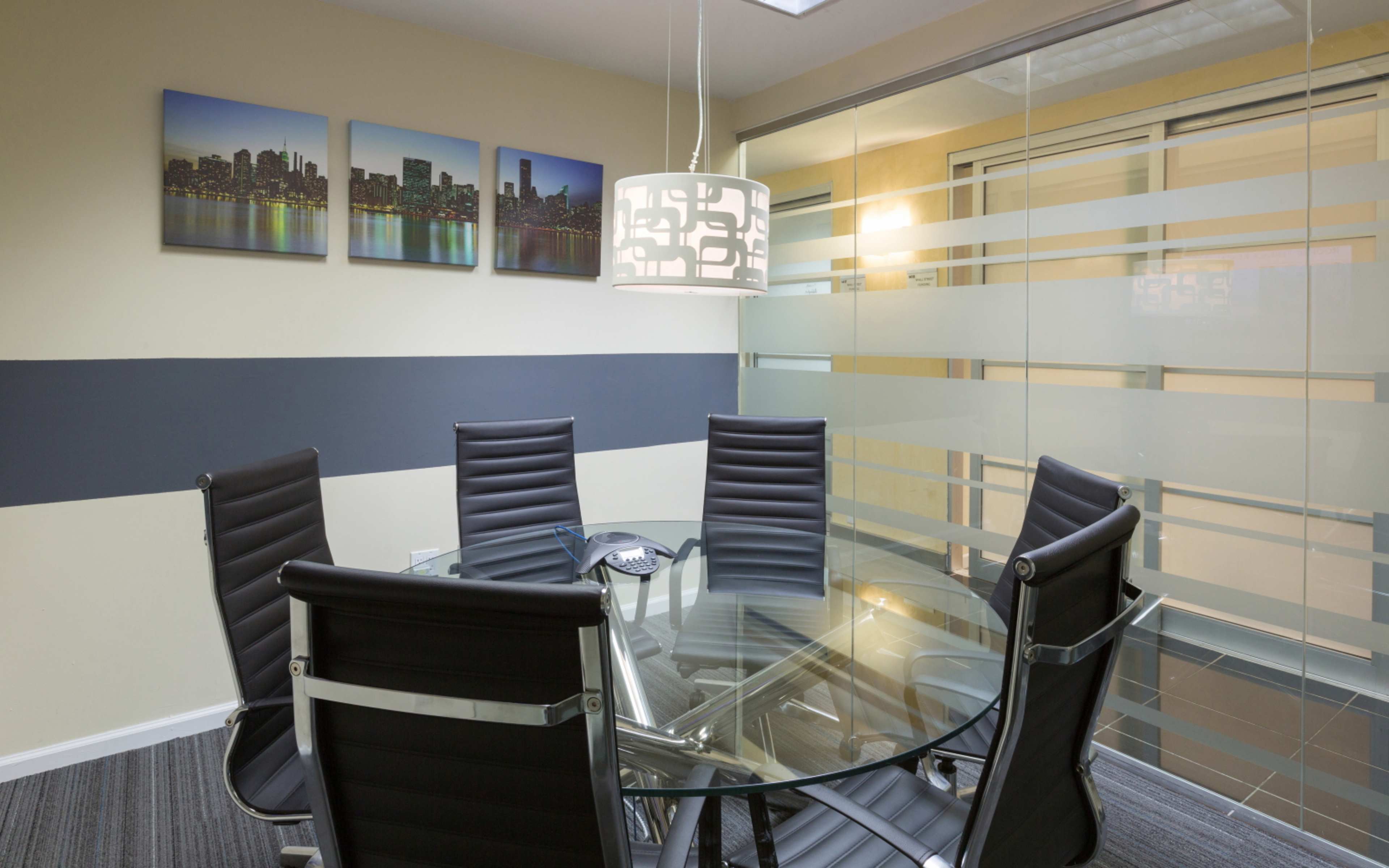 A modern conference room featuring a round glass table with six black leather chairs, surrounded by a wall displaying city skyline artwork and a pendant light.