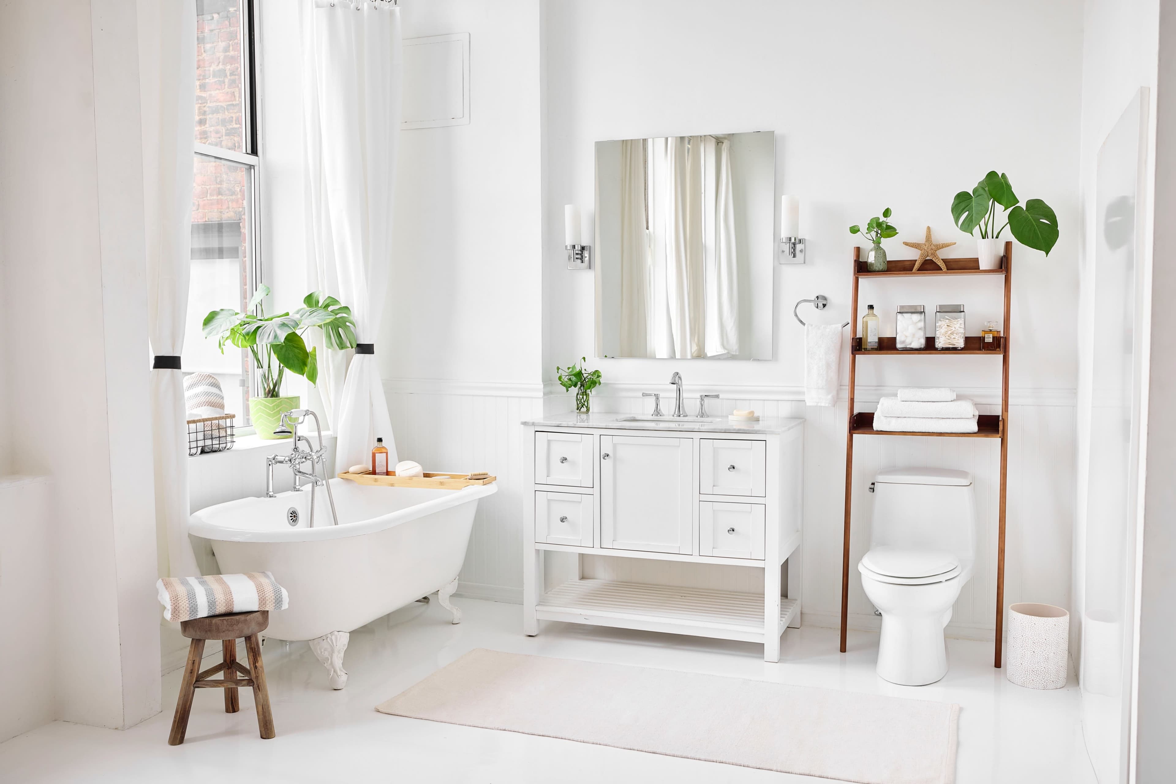 The image shows a bright, modern bathroom featuring a freestanding bathtub, a white vanity with a mirror, a wooden shelf with towels, and a toilet.