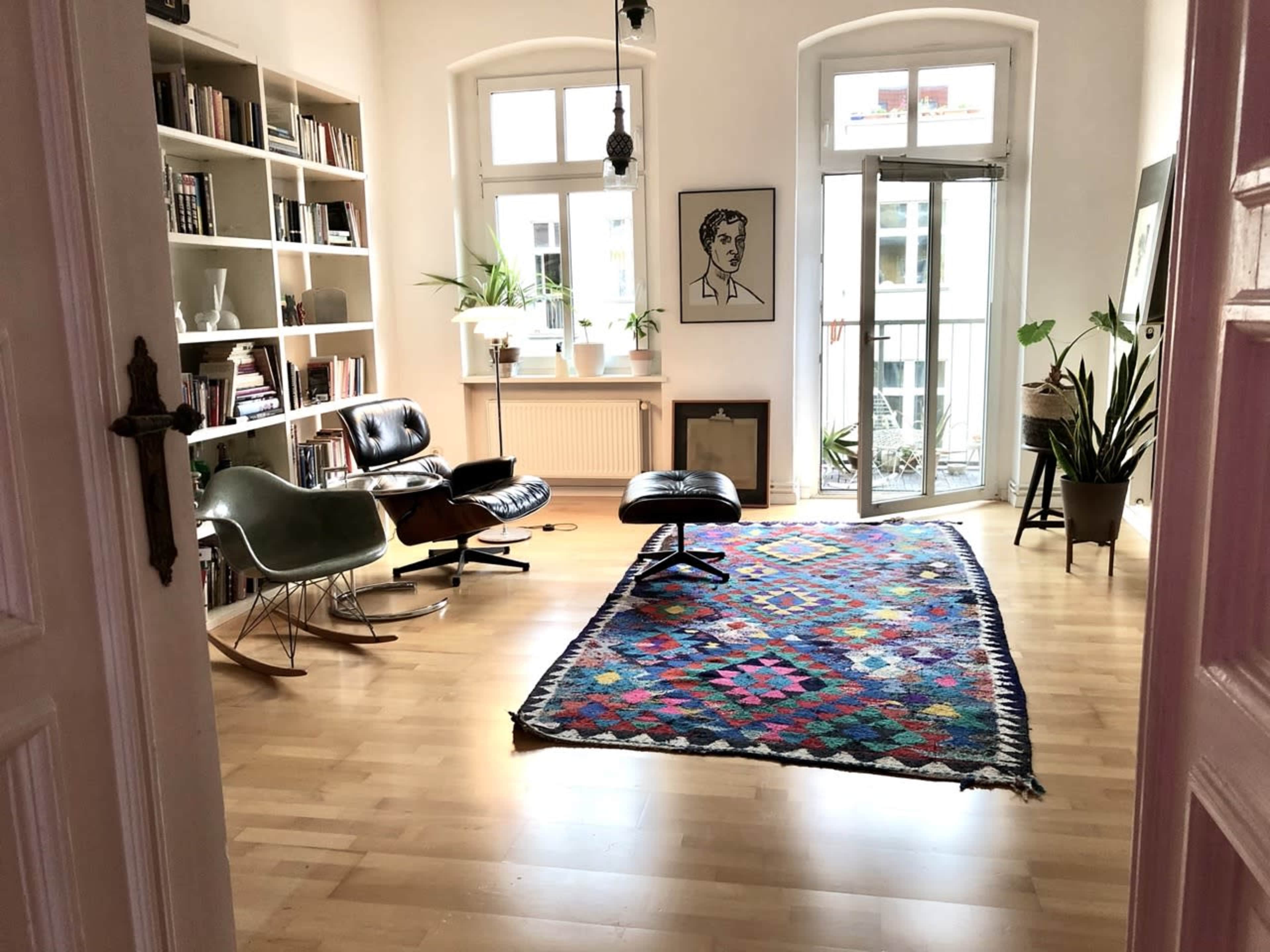 A brightly lit living room features a colorful rug, modern seating, bookshelves, and potted plants near large windows.
