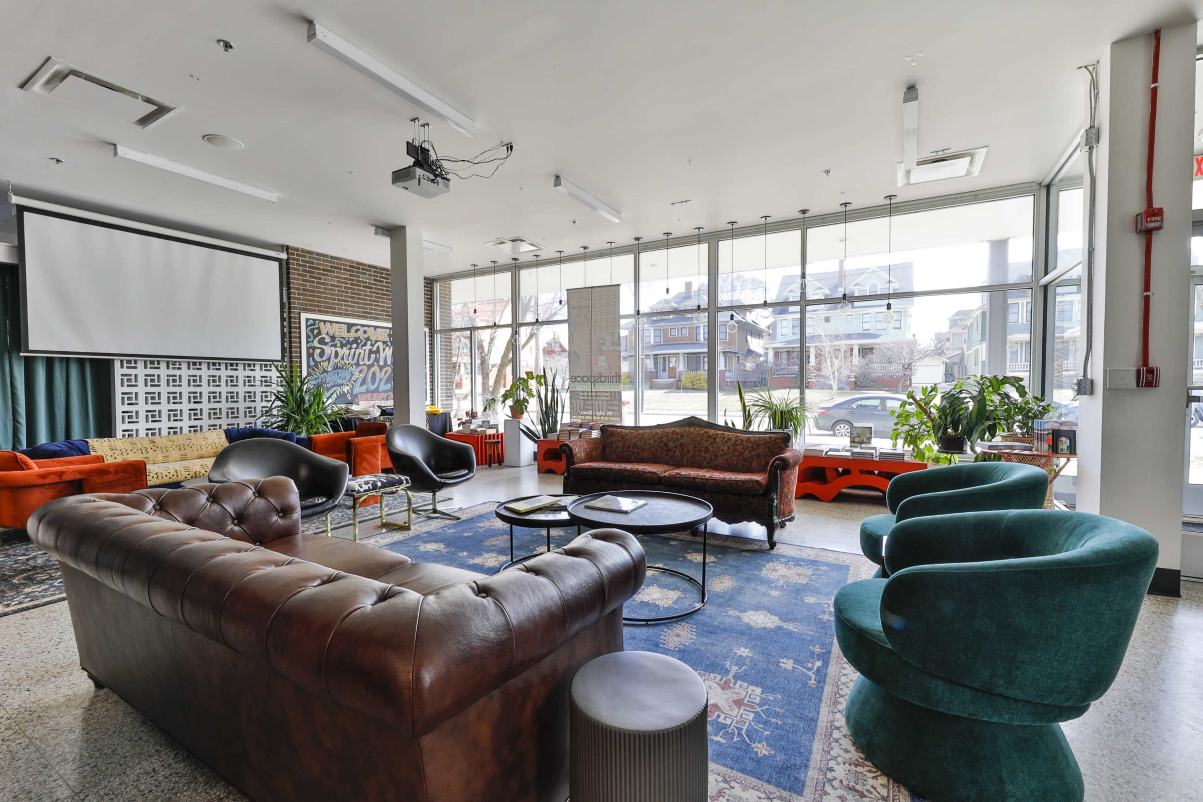 A modern lounge area with a mix of seating options, large windows allowing natural light, and various plants.