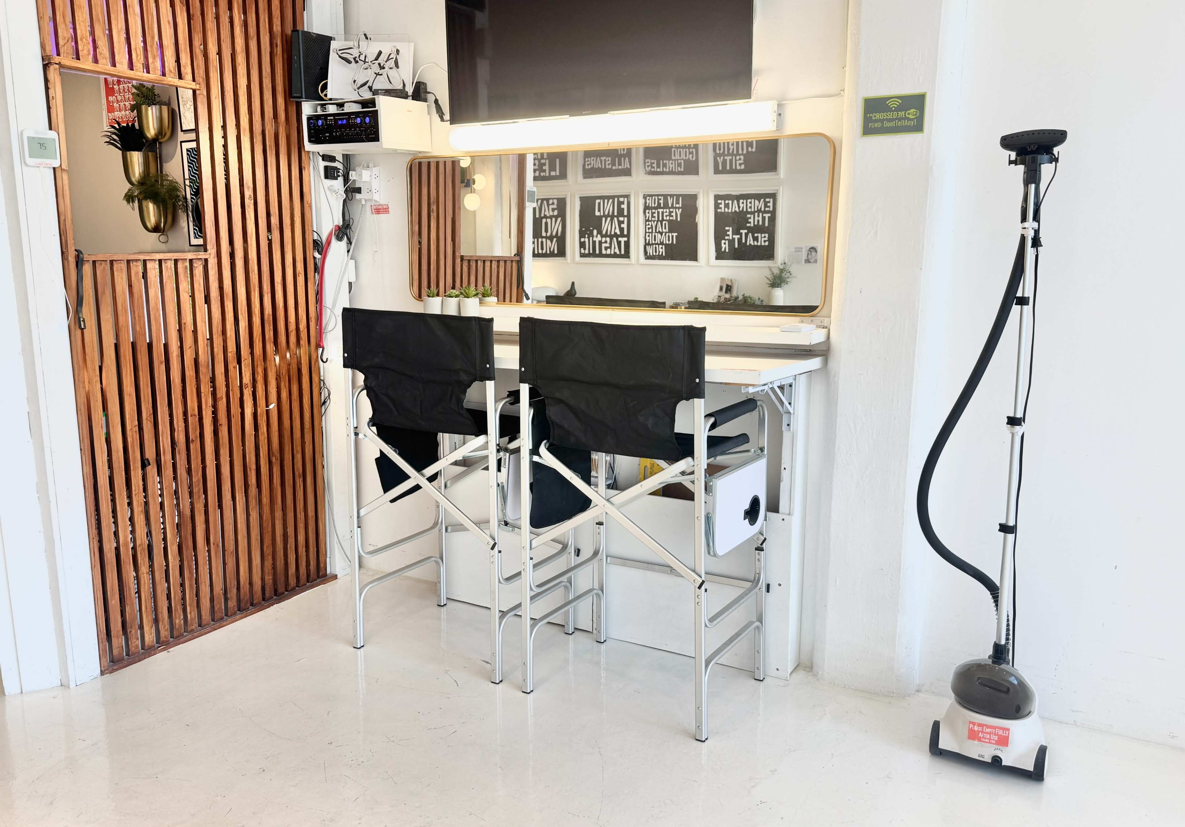 The image shows a modern, minimalist workspace featuring two black folding chairs at a table with a mirror and various items on the wall, alongside a clothes steamer in the corner.
