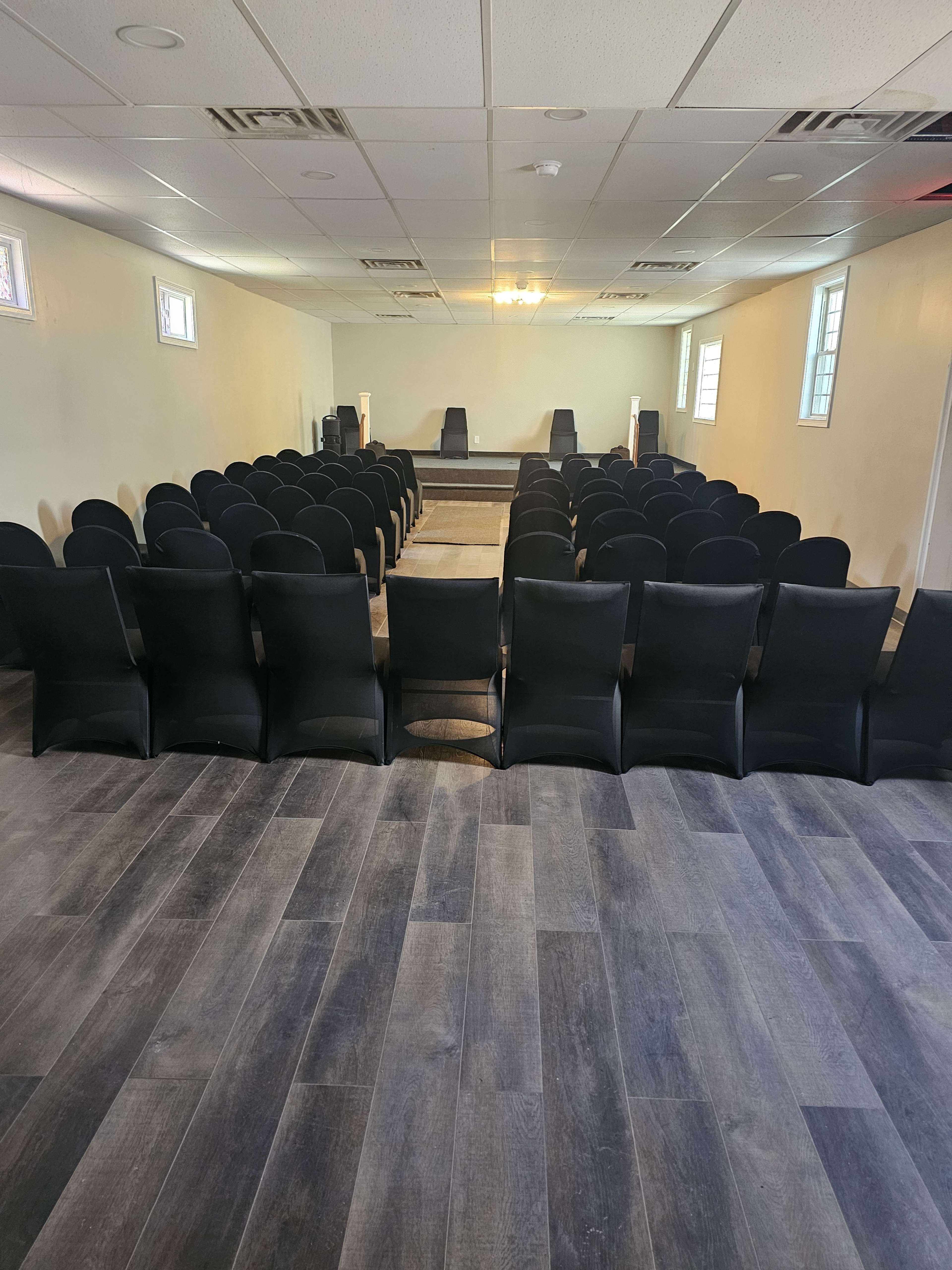 The image shows a spacious interior room arranged with rows of black chairs facing a small stage or platform at the front.