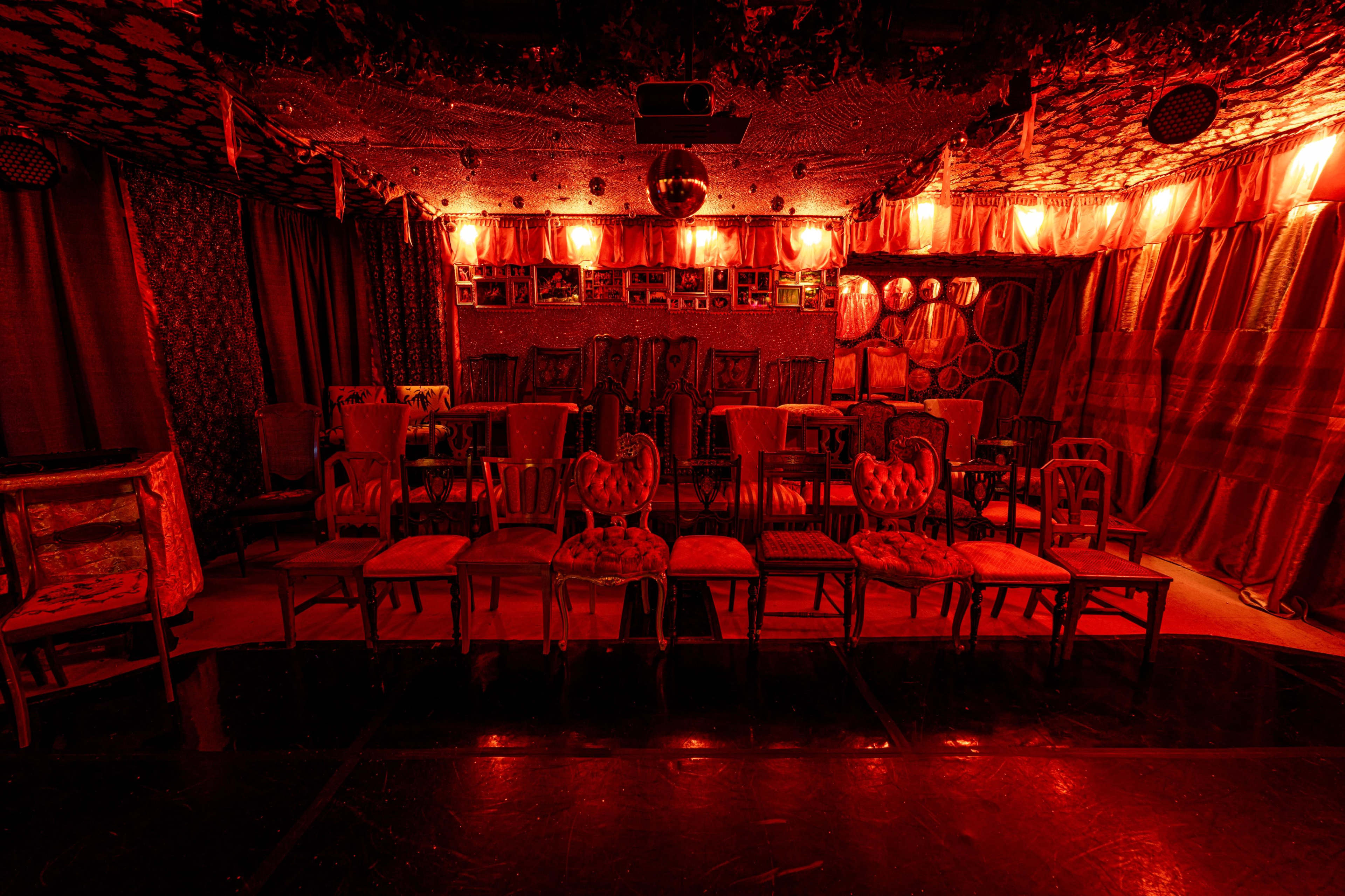 A dimly lit room features rows of vintage chairs under red lights, with patterned walls and decorative elements.