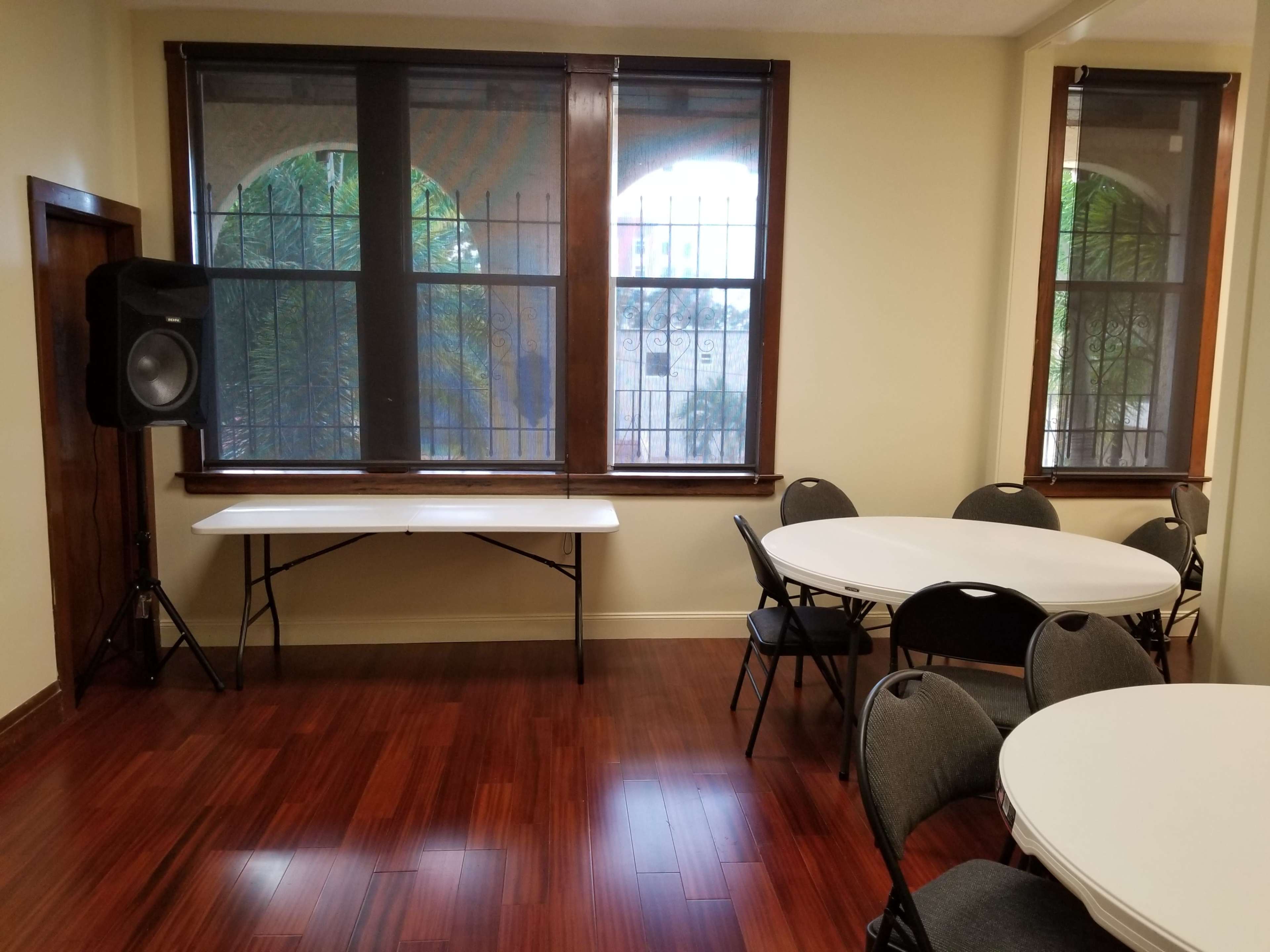 A room with wooden flooring, two round tables with chairs, a folding table, and a speaker next to the windows.